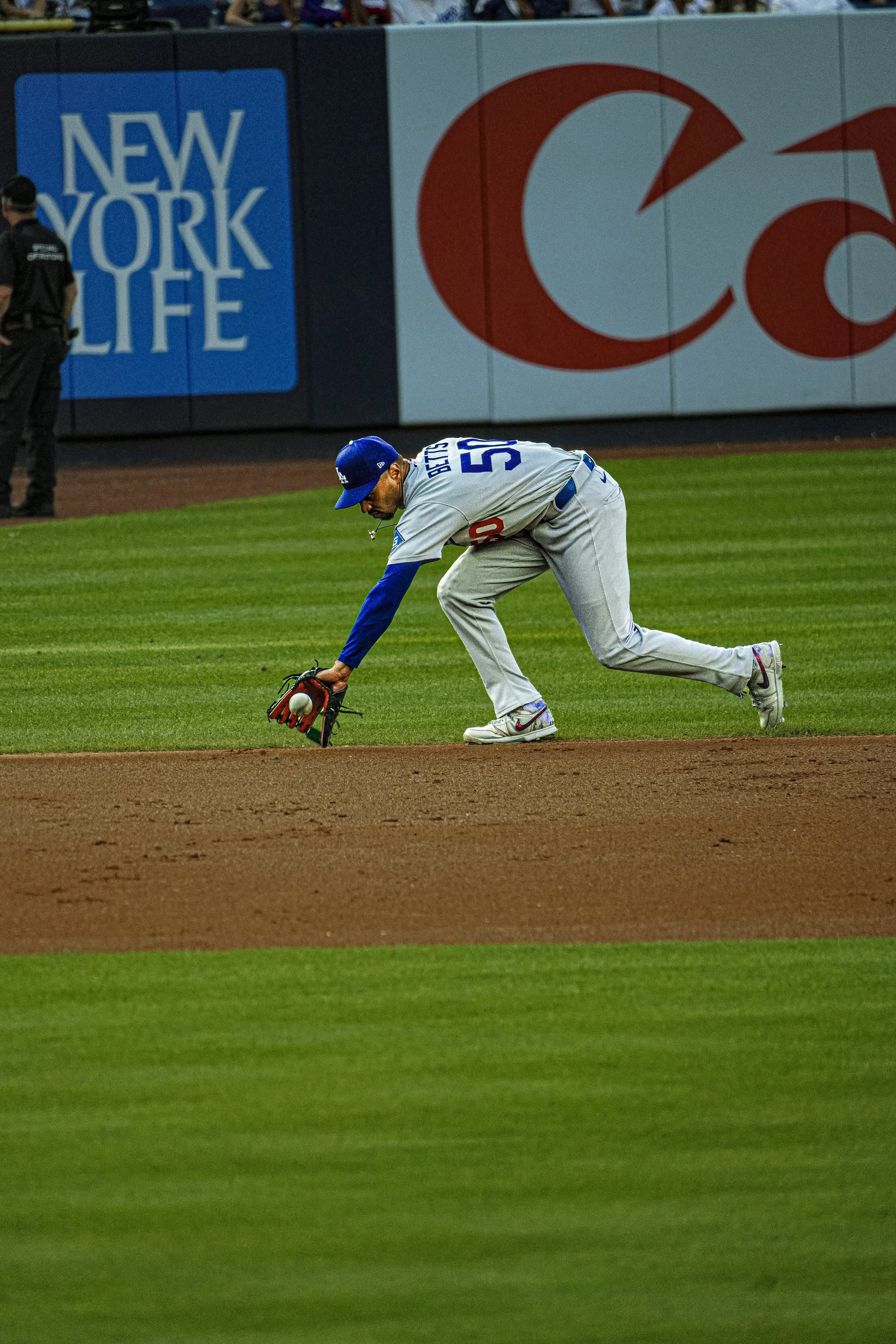 A baseball player in a gray uniform with blue accents is crouching on the field, reaching for a ball near the ground. The player appears to be a fielder attempting to stop the ball. There are advertisements on the outfield wall, including a blue sign