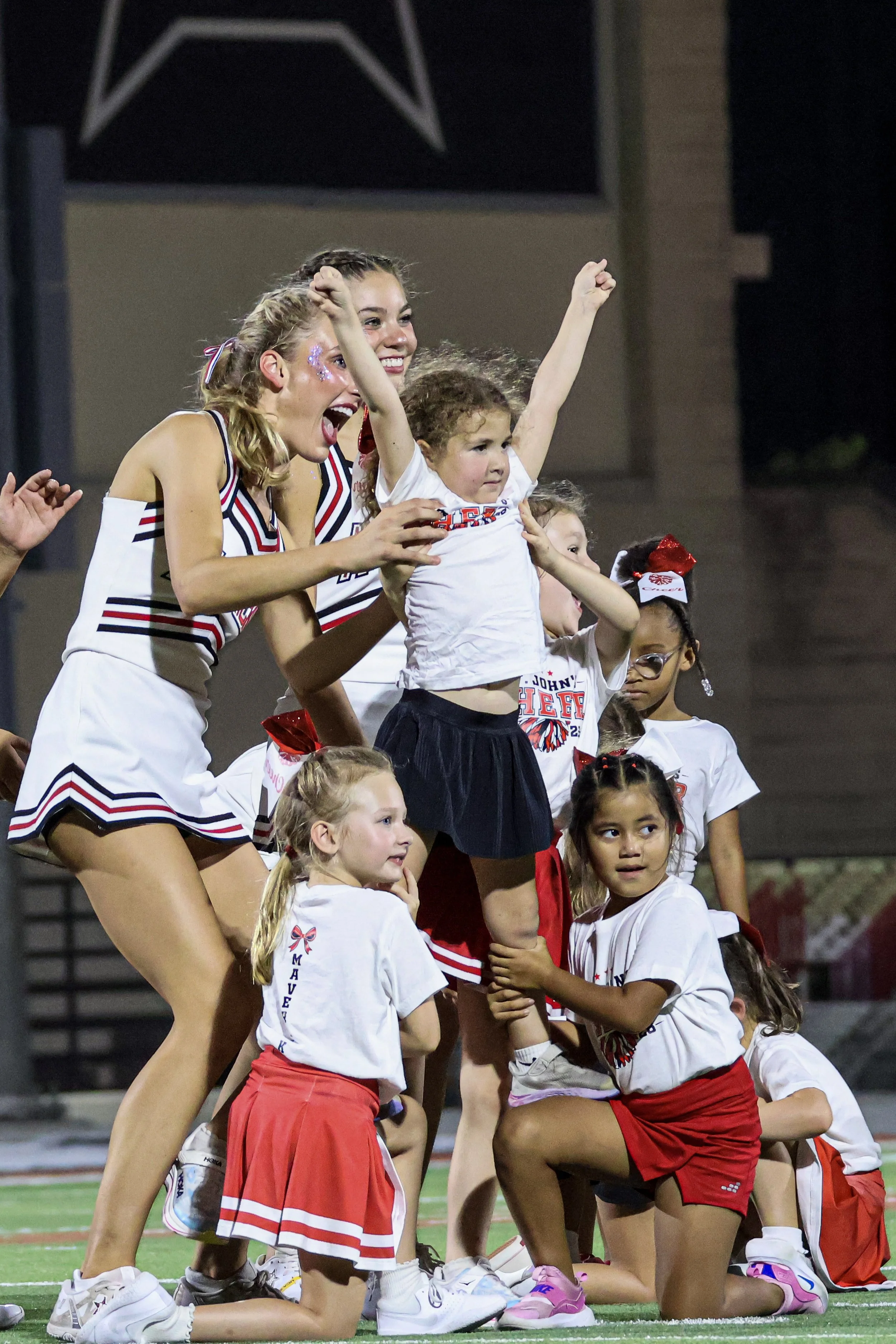 Children and young women cheer on a football field, celebrating during a game or event at night. They are wearing sports uniforms and appear to be happy and excited.