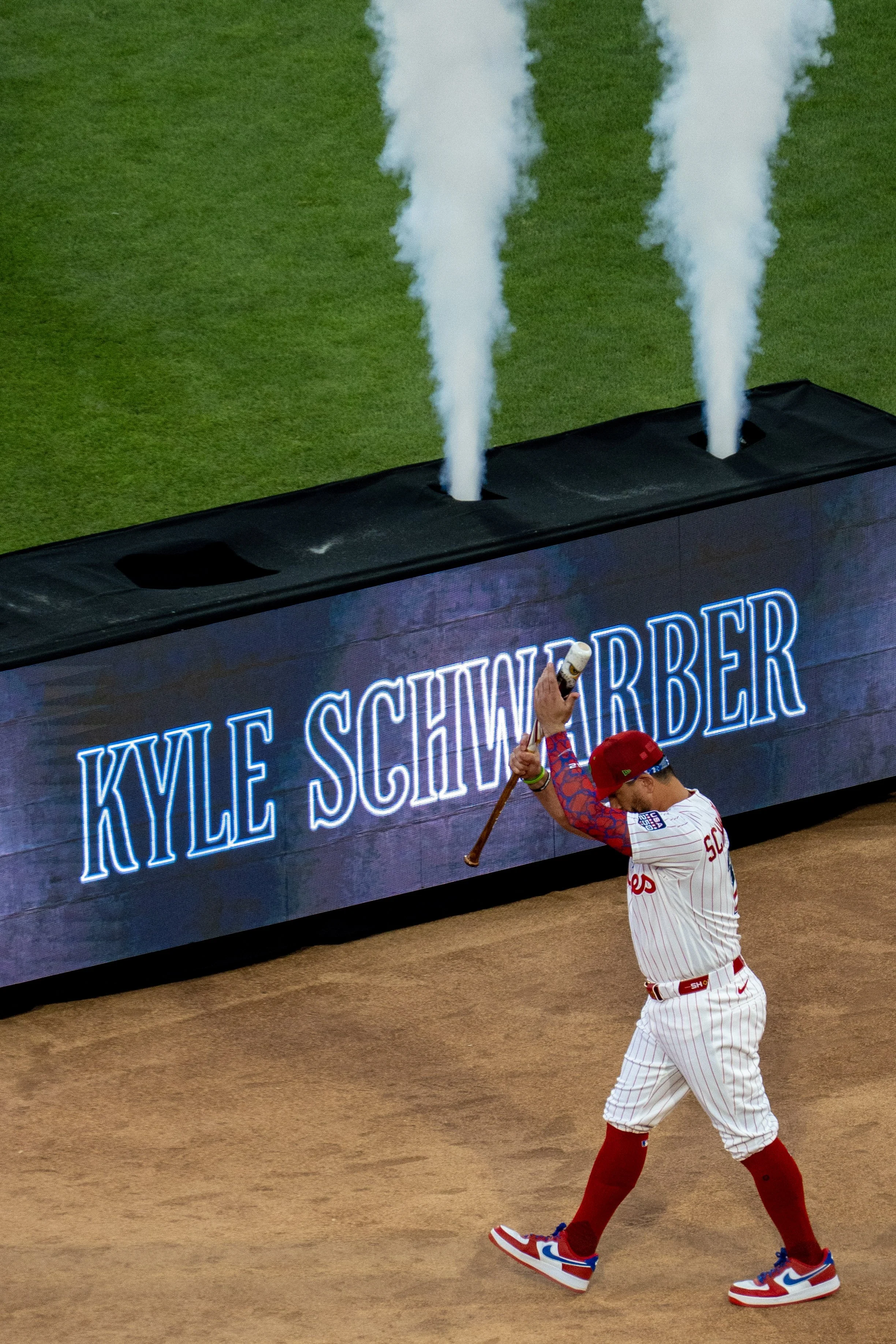 A baseball player in a white pinstripe uniform with red accents is standing on the field with his head bowed. He is wearing blue, white, and red sneakers and red socks, with a red hat. In the background, a digital scoreboard displays the name "KYLE S