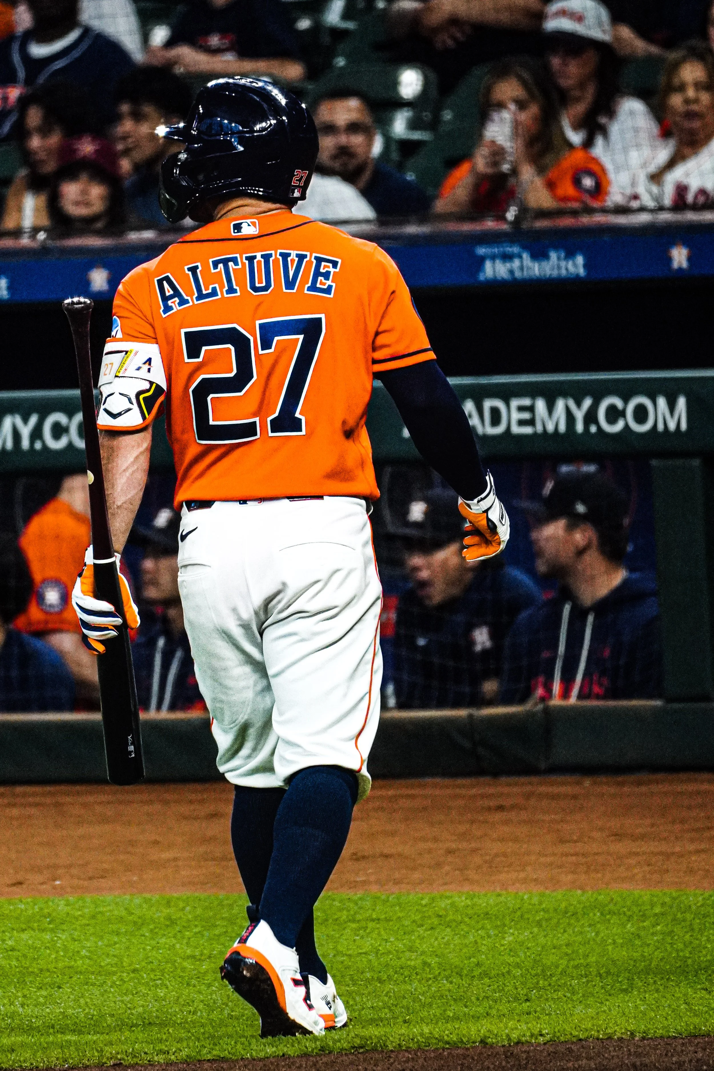 A baseball player in a bright orange uniform with the name "ALTUVE" and the number 27 on the back, walking on the field while holding a bat, with spectators watching from the stands.