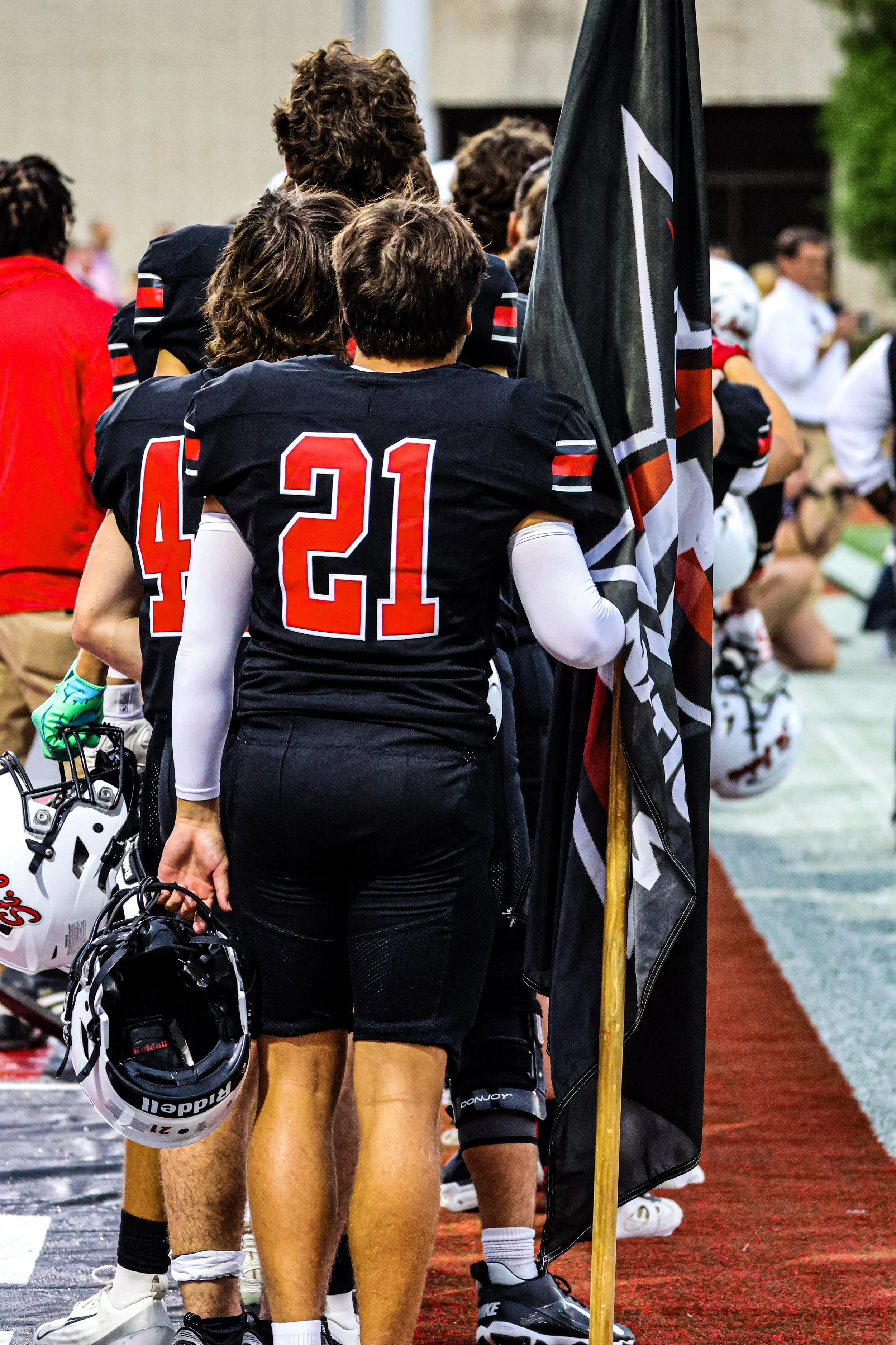 American football players wearing black jerseys with red and white accents, standing on the sideline with helmets in hand, in a stadium with a football field in the background.