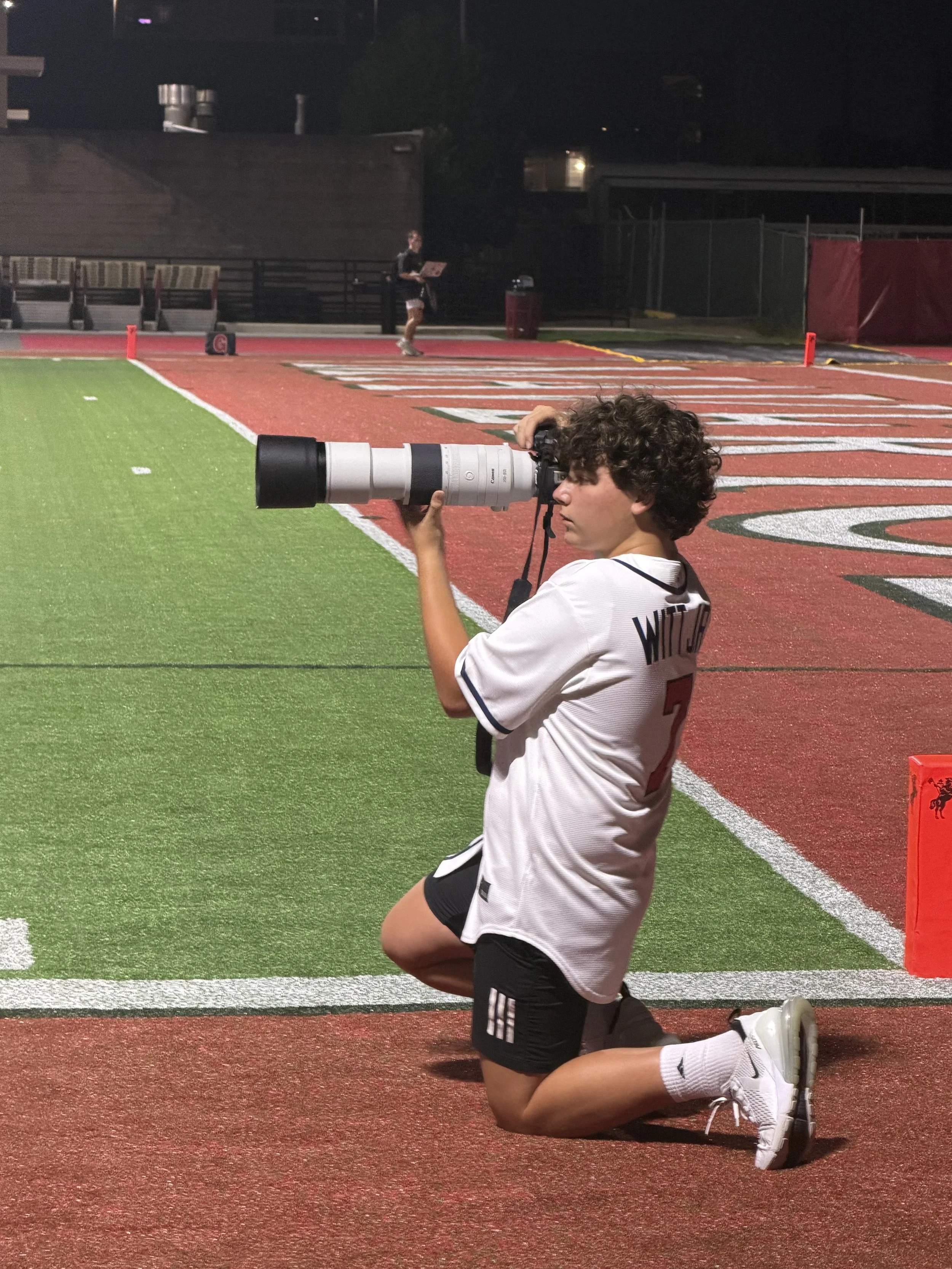 A young man in a sports uniform kneels on one knee on a sports field, holding a large camera with a long telephoto lens, aiming it towards the front, near the sideline at night. There is another person in the background standing near a fence with a laptop.