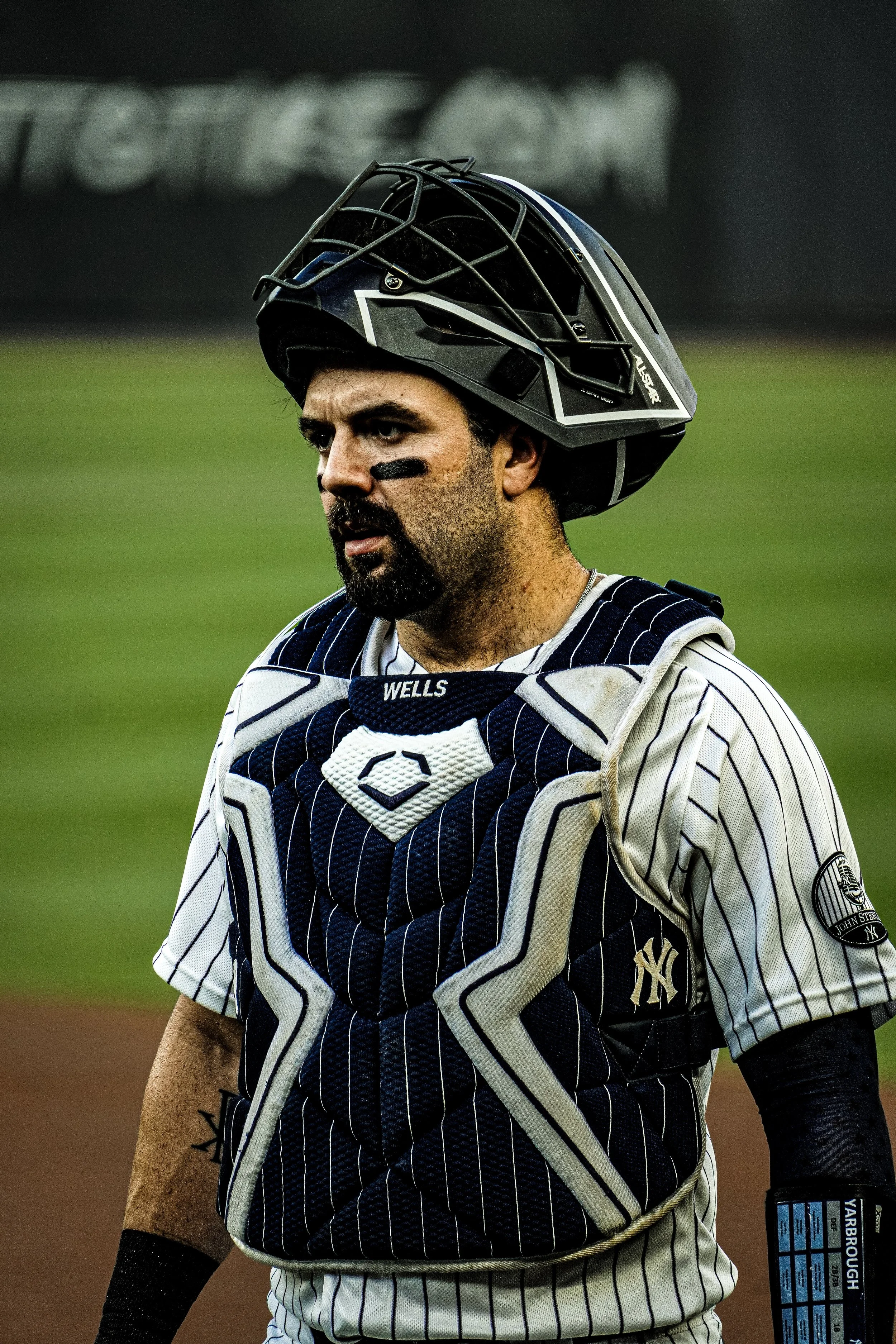 A baseball player in a pinstriped uniform and a protective helmet with a faceguard on a baseball field. The player has a beard and black eye paint under his eyes. He is also wearing a chest protector with the New York Yankees logo.