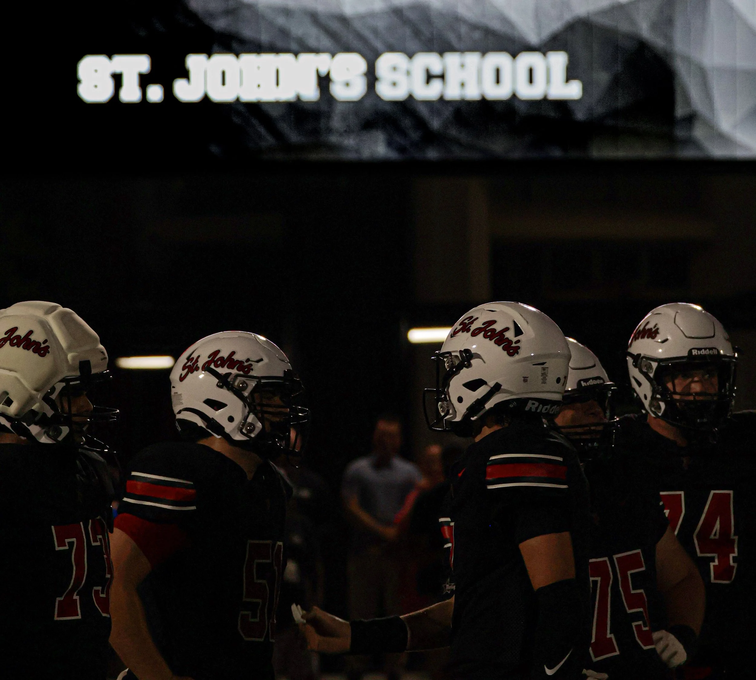 A group of football players wearing black jerseys and white helmets with 'St. John’s' logo, standing in formation inside a stadium with a sign overhead reading 'St. John's School'.