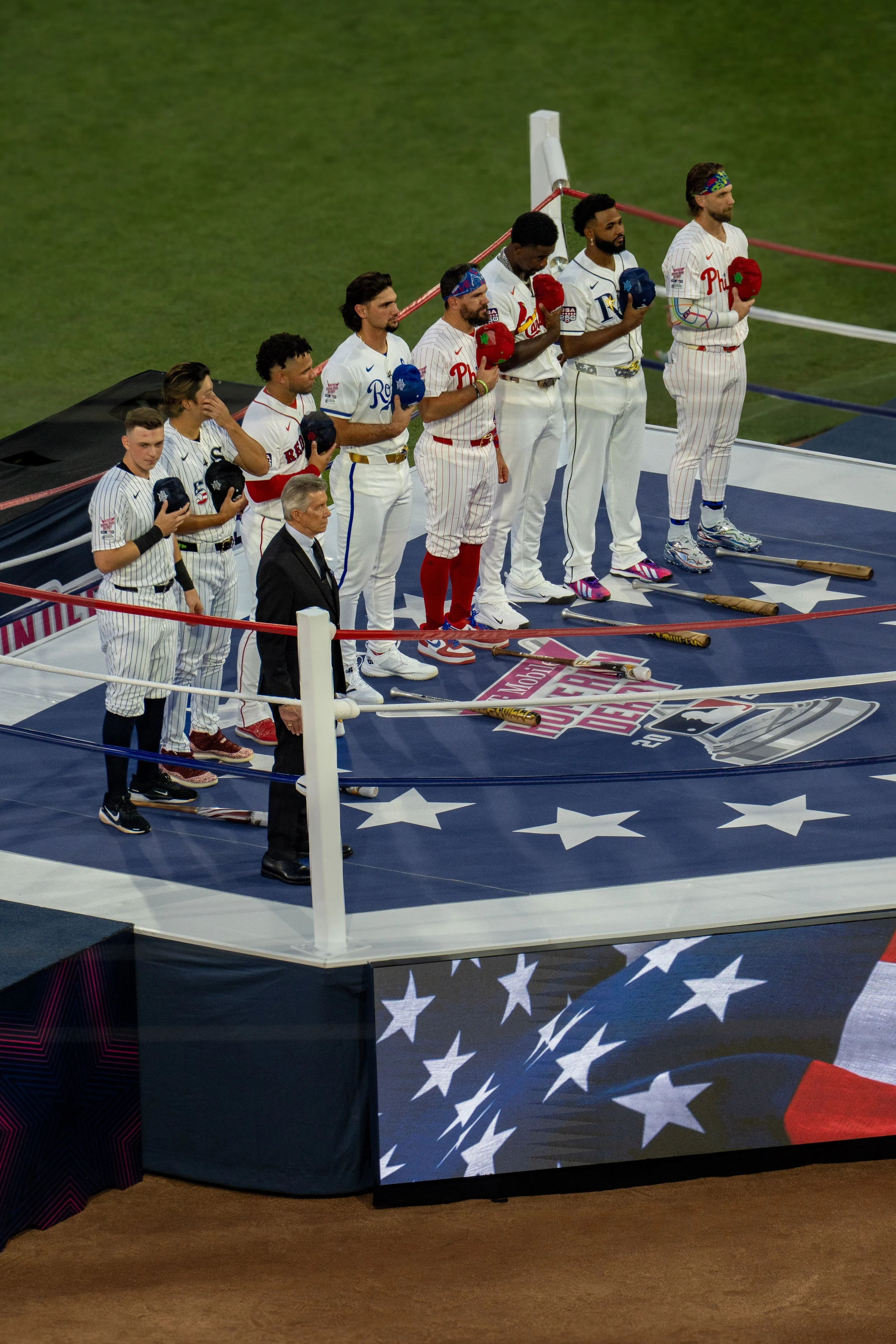 Men in baseball uniforms standing on a platform during the national anthem, holding caps over their hearts, with a baseball bat on the ground in front of them, on a field decorated with American flag stars.