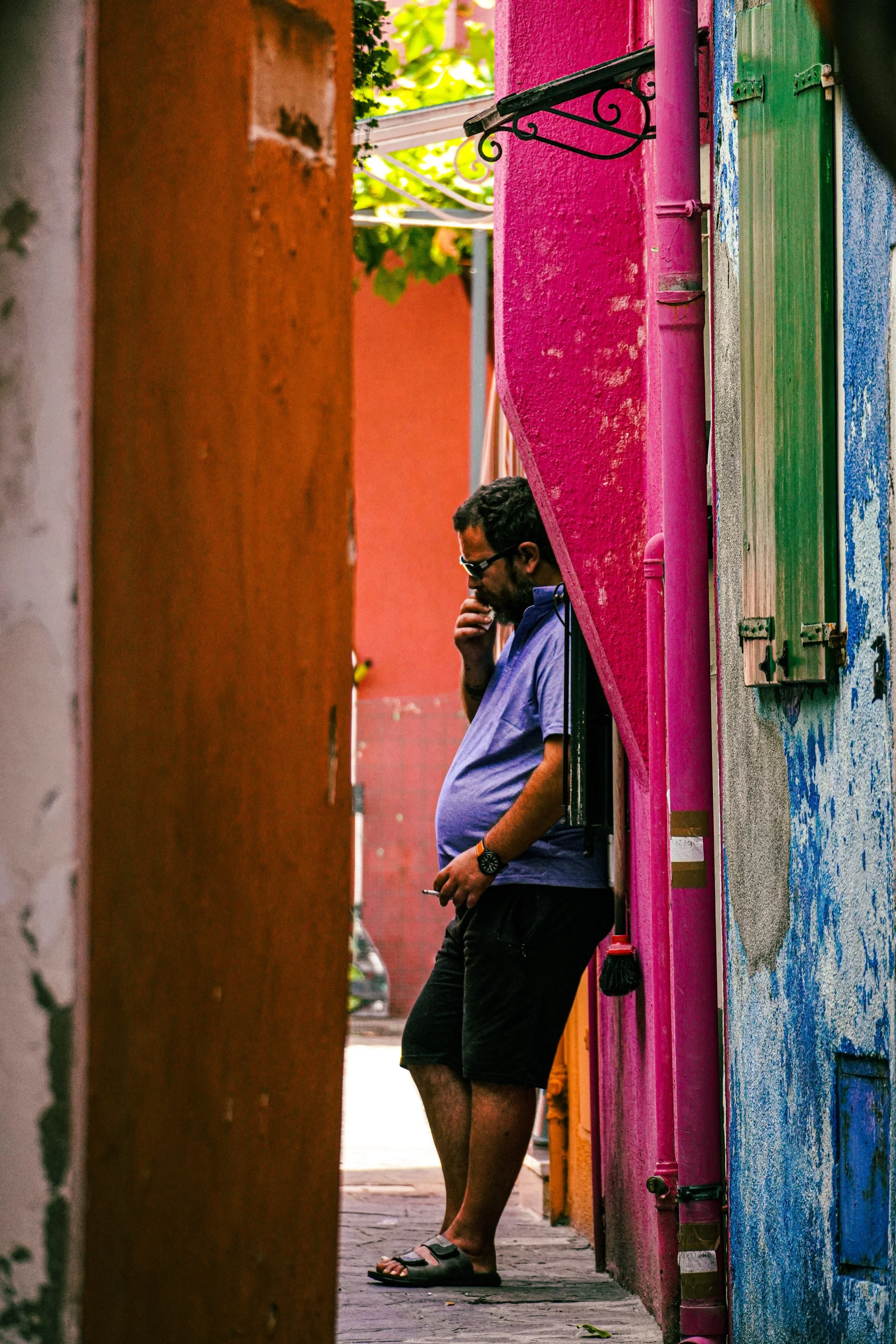 A man wearing glasses, a blue shirt, and black shorts is standing with his back against a vibrant pink and green wall, holding a cigarette near his mouth and looking down. The scene is colorful with textured walls and a narrow alleyway, with hanging 