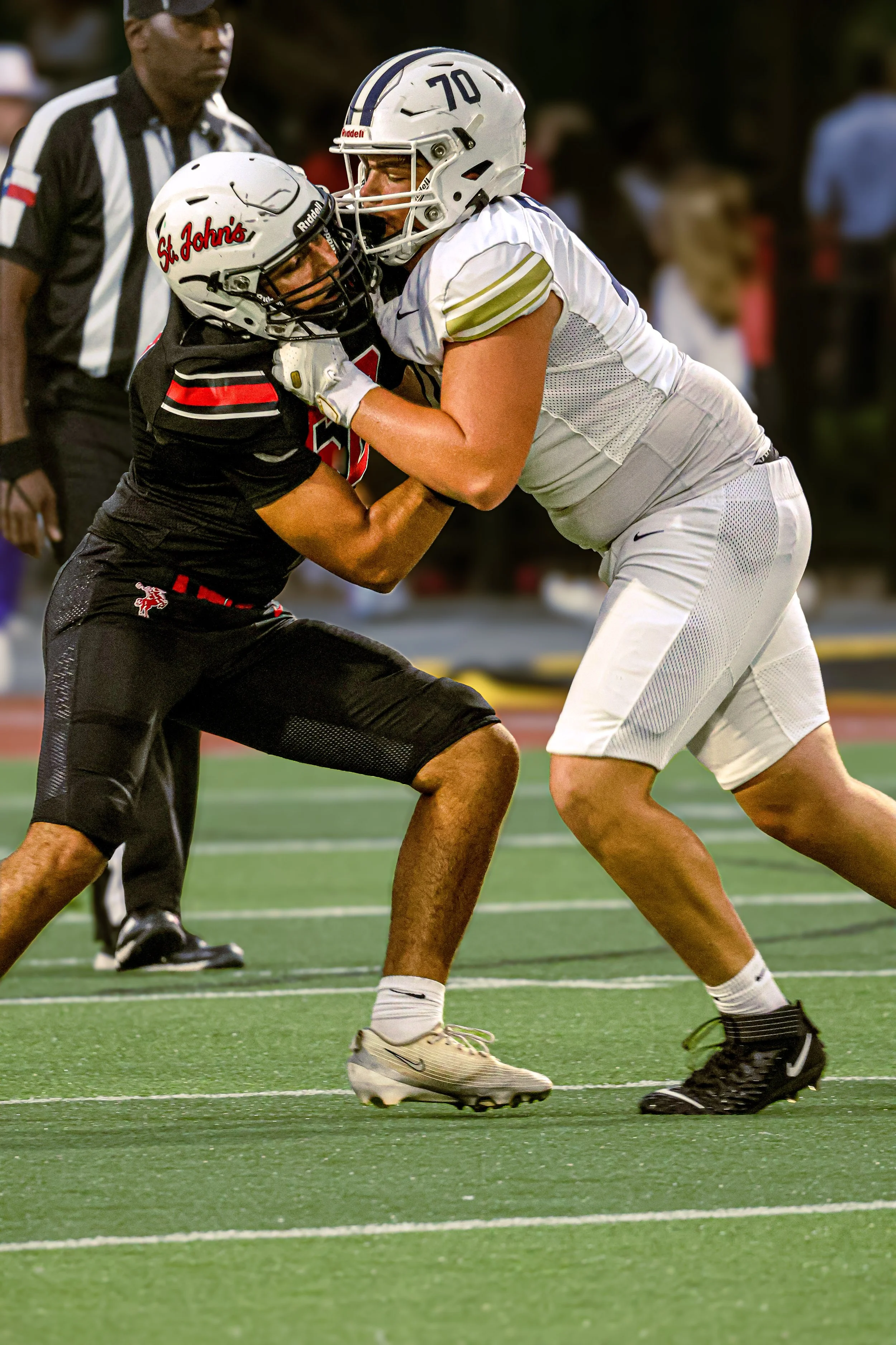 Two football players are engaged in a tackle on a football field. One player, wearing a black uniform with a helmet labeled 'St. John's,' is being protected by the other player, in a white uniform and helmet. An official referee stands in the backgro