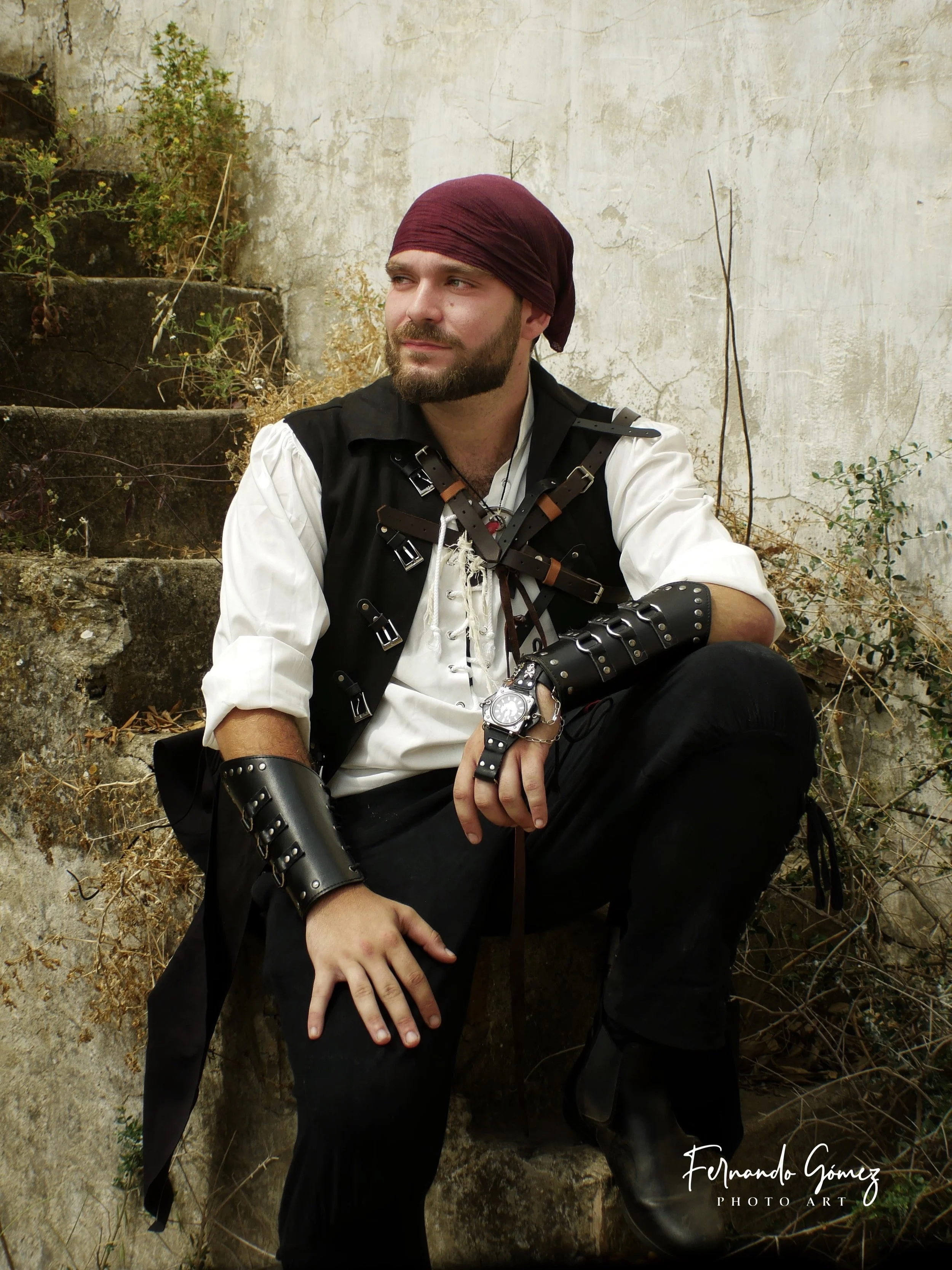 A man sitting outdoors against a weathered wall with plants and stairs in the background. He has a beard, wears a purple headscarf, black leather armor with buckles, a white shirt with rolled sleeves, and black pants. He has a wristwatch and leather 