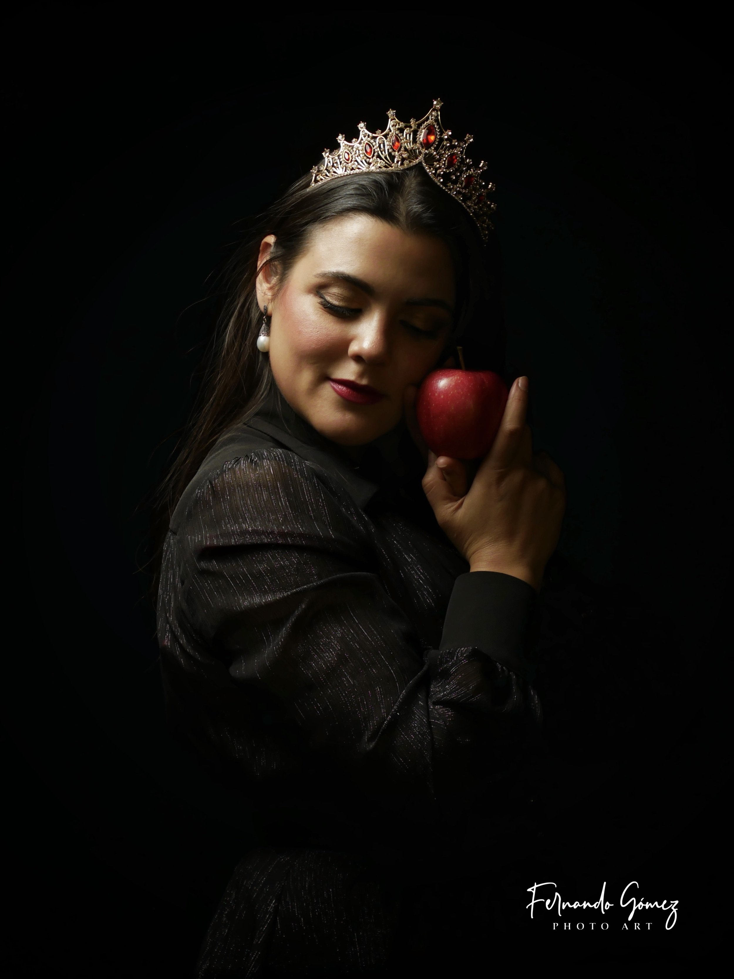 A woman with closed eyes holding a red apple, wearing a tiara and pearl earrings, set against a black background.