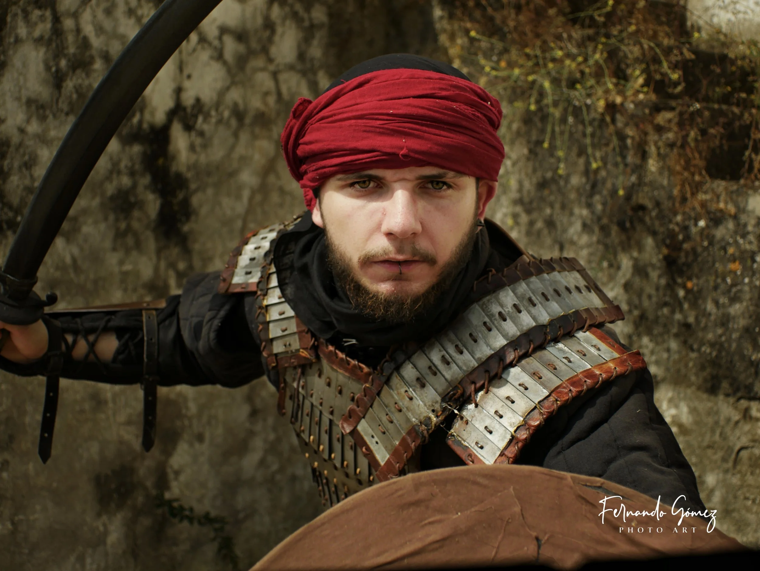 A man wearing a red headwrap and armor made of metal plates, standing outdoors against a rocky background.