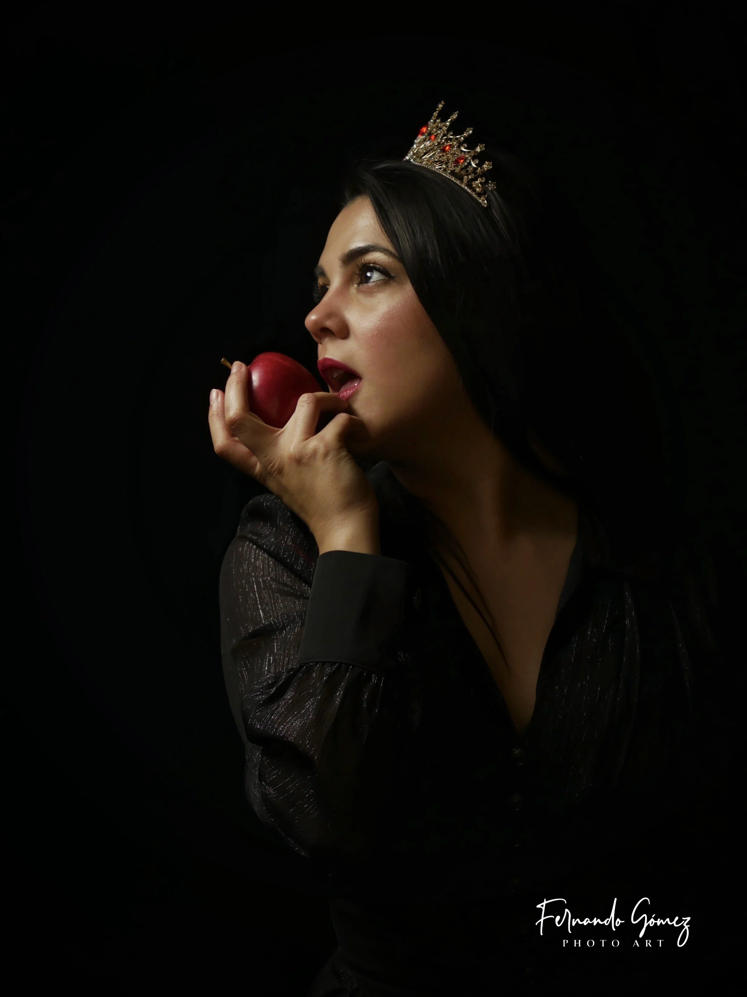 A woman with dark hair and light skin wearing a small crown, holding a red apple near her lips, against a black background. She appears to be looking upwards. Her makeup is subtle and she is wearing a black, shiny, long-sleeved garment.