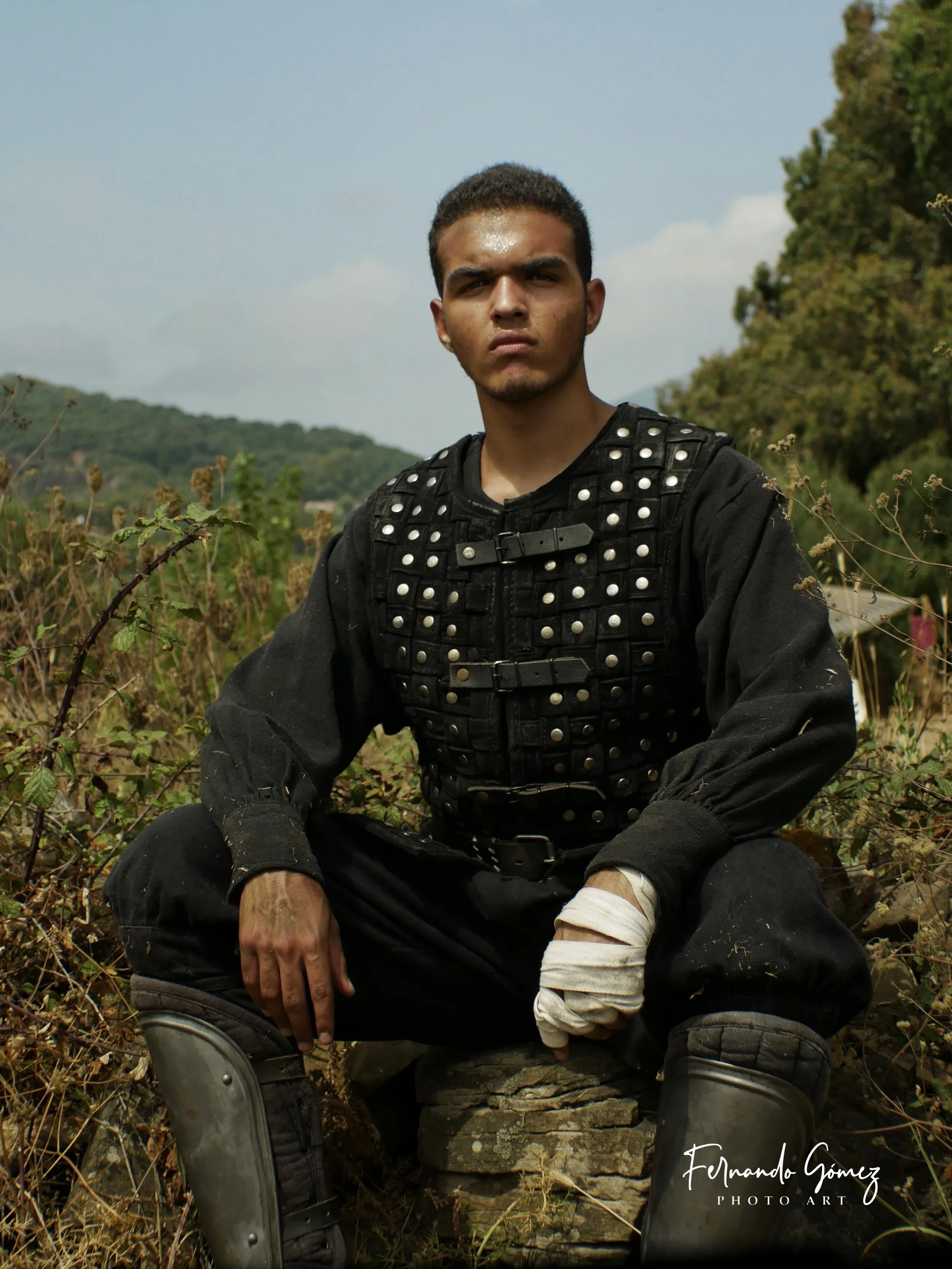 Young man with short dark hair and tan skin sitting outdoors in a natural setting, wearing a black vest with metal studs and black pants, with a bandage on his right ankle. He has a serious expression and is positioned in front of a landscape with gr