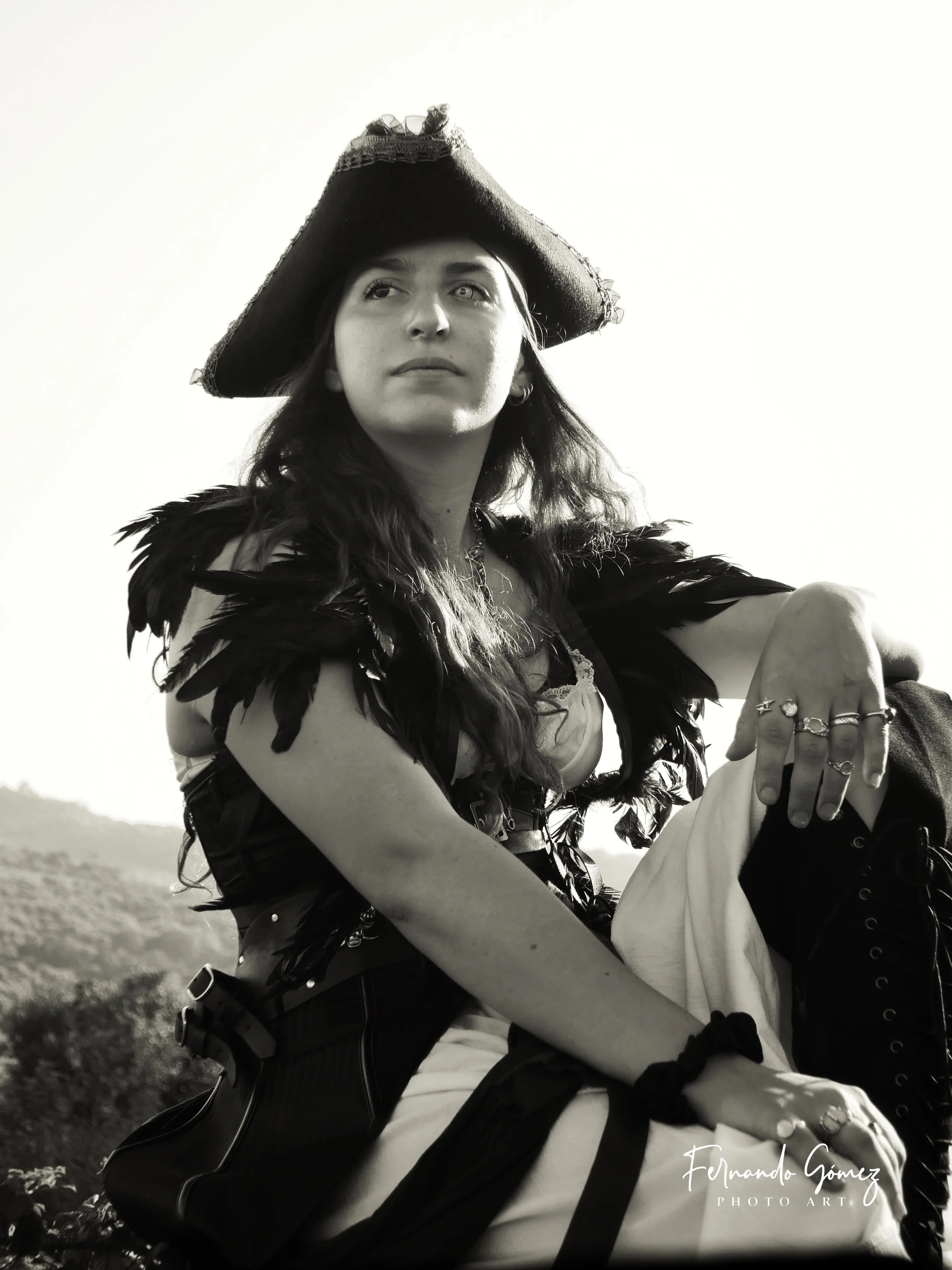 Black and white photograph of a woman dressed as a pirate, wearing a large pirate hat with frayed edges, feathered clothing, and jewelry, sitting outdoors with a blurred natural landscape in the background.