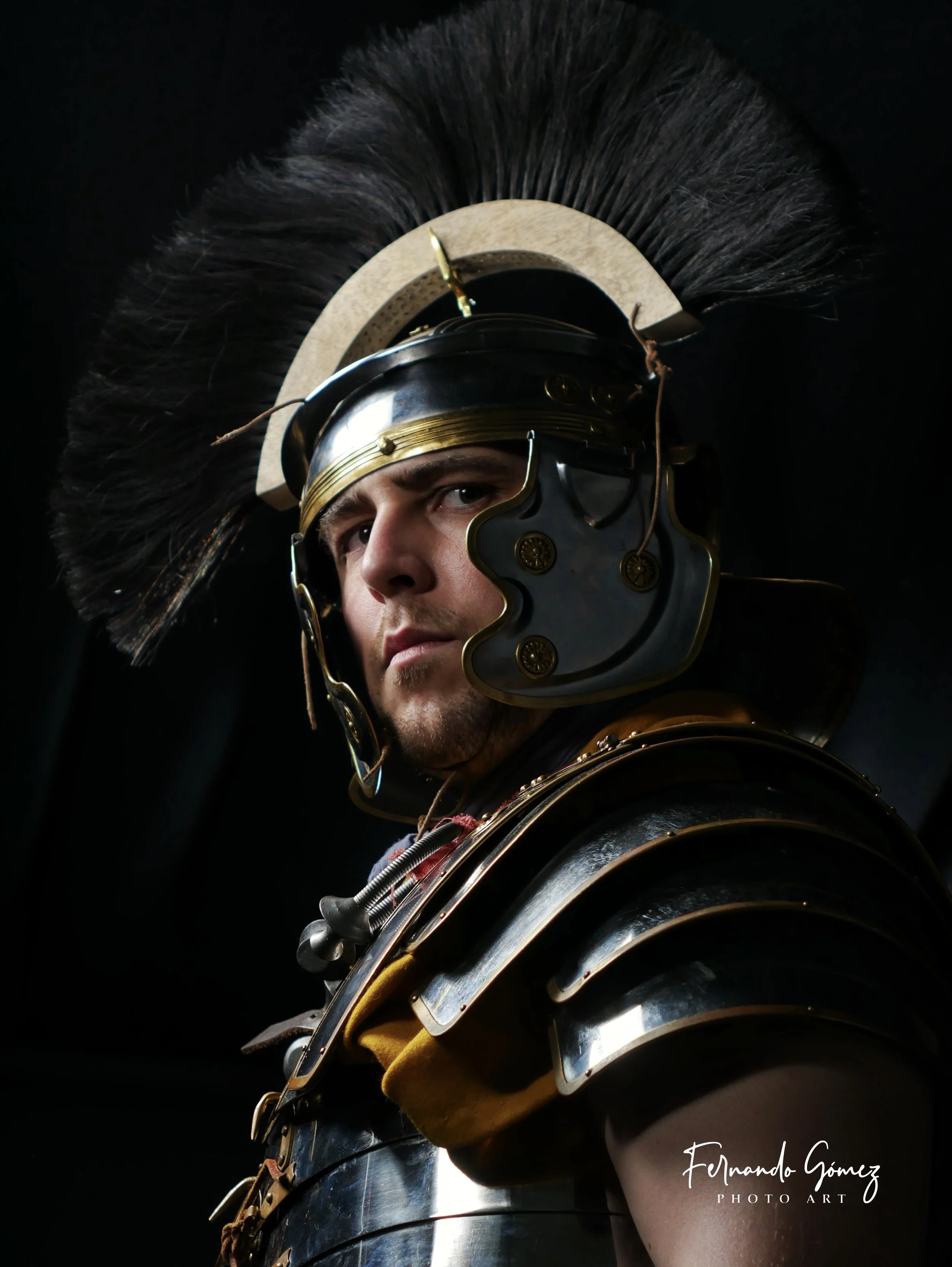 A person dressed as a Roman soldier wearing a metallic helmet with an elaborate crest of black horsehair, a leather neck guard, and metallic armor with gold and silver accents, set against a black background.