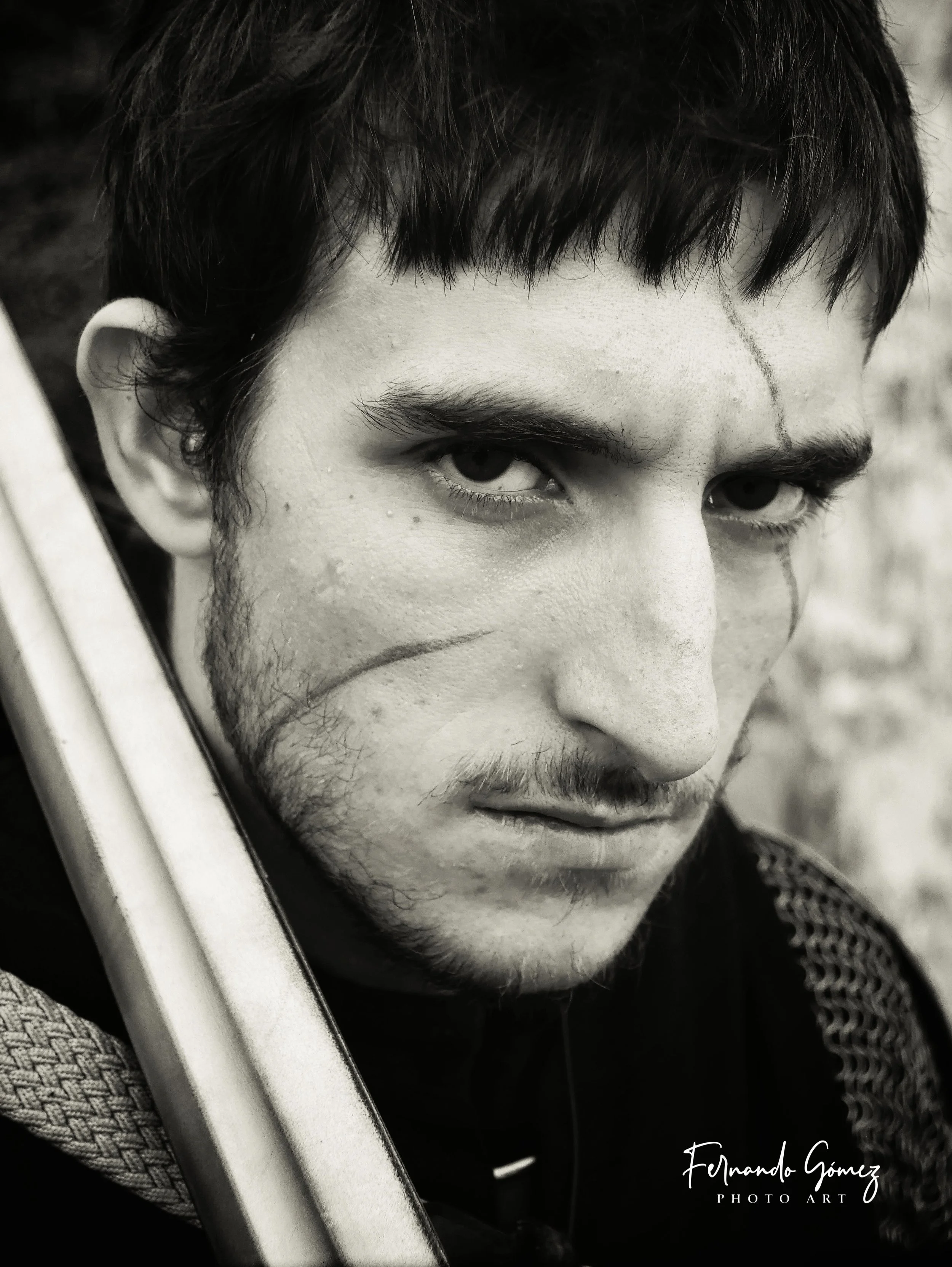 Black-and-white close-up photograph of a man's face showing intense eyes, dark hair, and facial hair, with a slightly reflective surface in the foreground holding a sword.