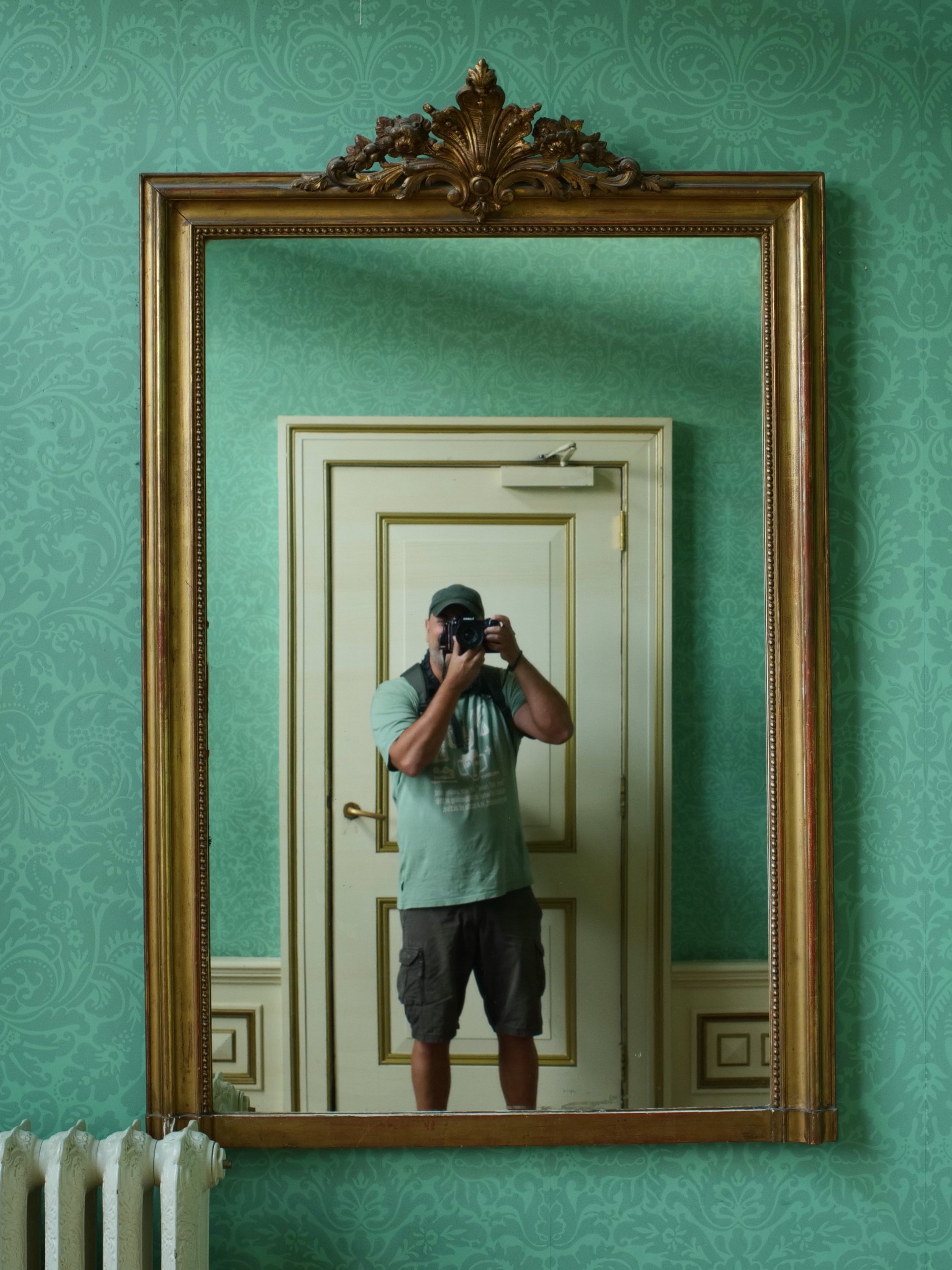 A large ornate gold-framed mirror on a teal patterned wall reflecting a person taking a photo of themselves. The person is wearing a baseball cap, green t-shirt, and shorts, standing in front of a white door with gold trim with their face partially obscured by the camera.