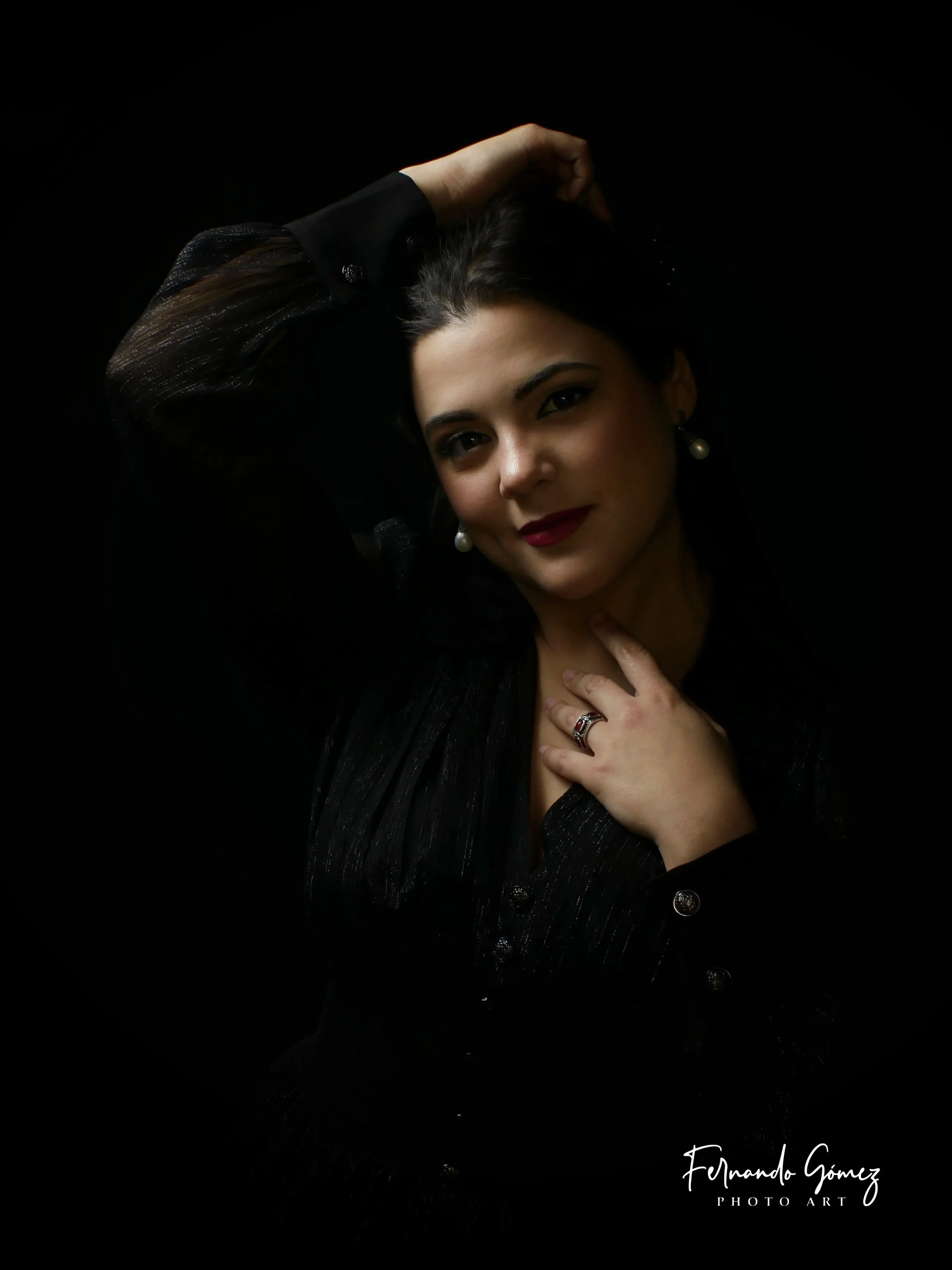 A portrait of a woman with dark hair, wearing pearl earrings and a black outfit with shiny textured fabric, posed with one hand touching her neck and the other behind her head, against a black background.