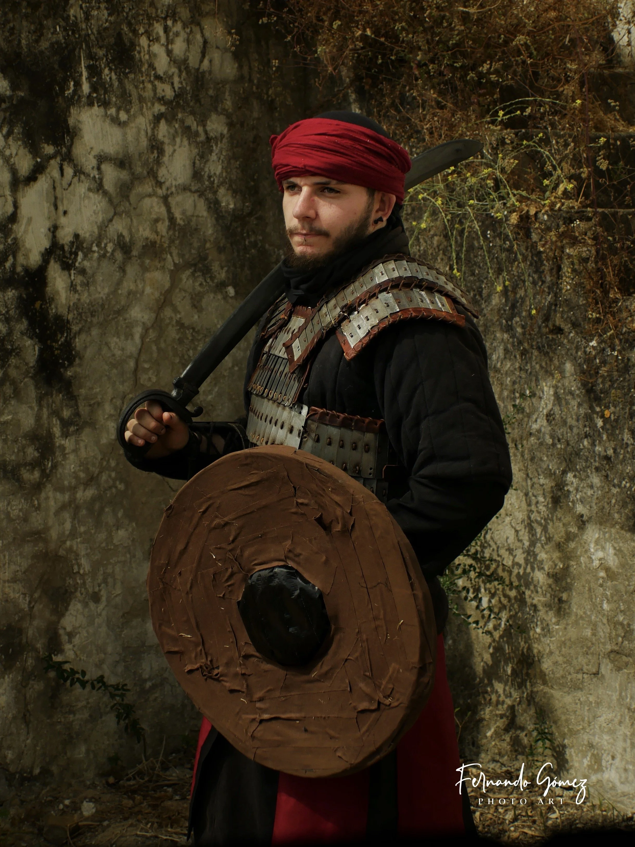 A man dressed as a warrior with a red headscarf, holding a sword on his shoulder, standing outdoors against a rocky background.