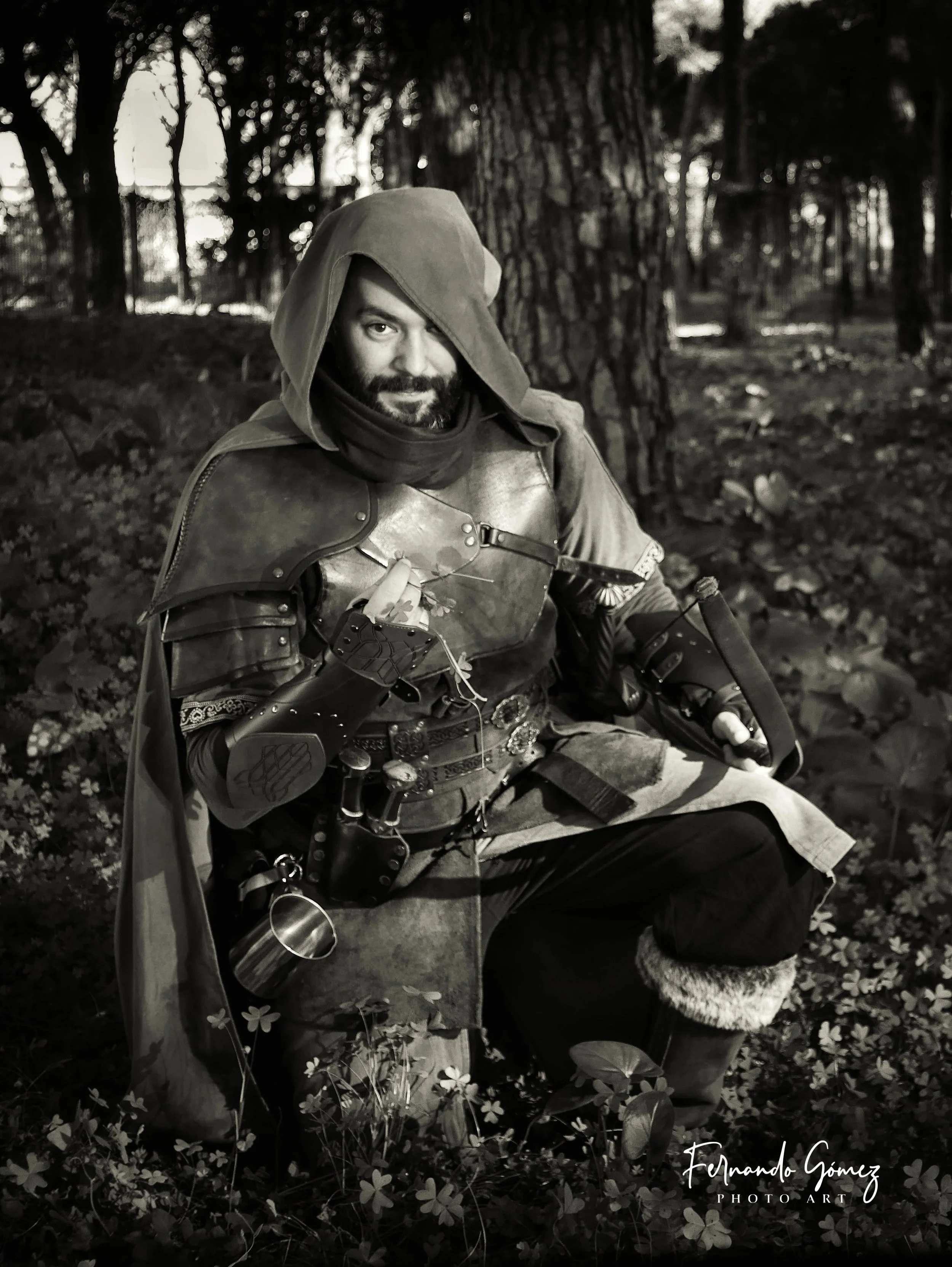 Black and white photo of a man wearing medieval armor and a hood, sitting outdoors among trees and flowers, with a confident expression. 