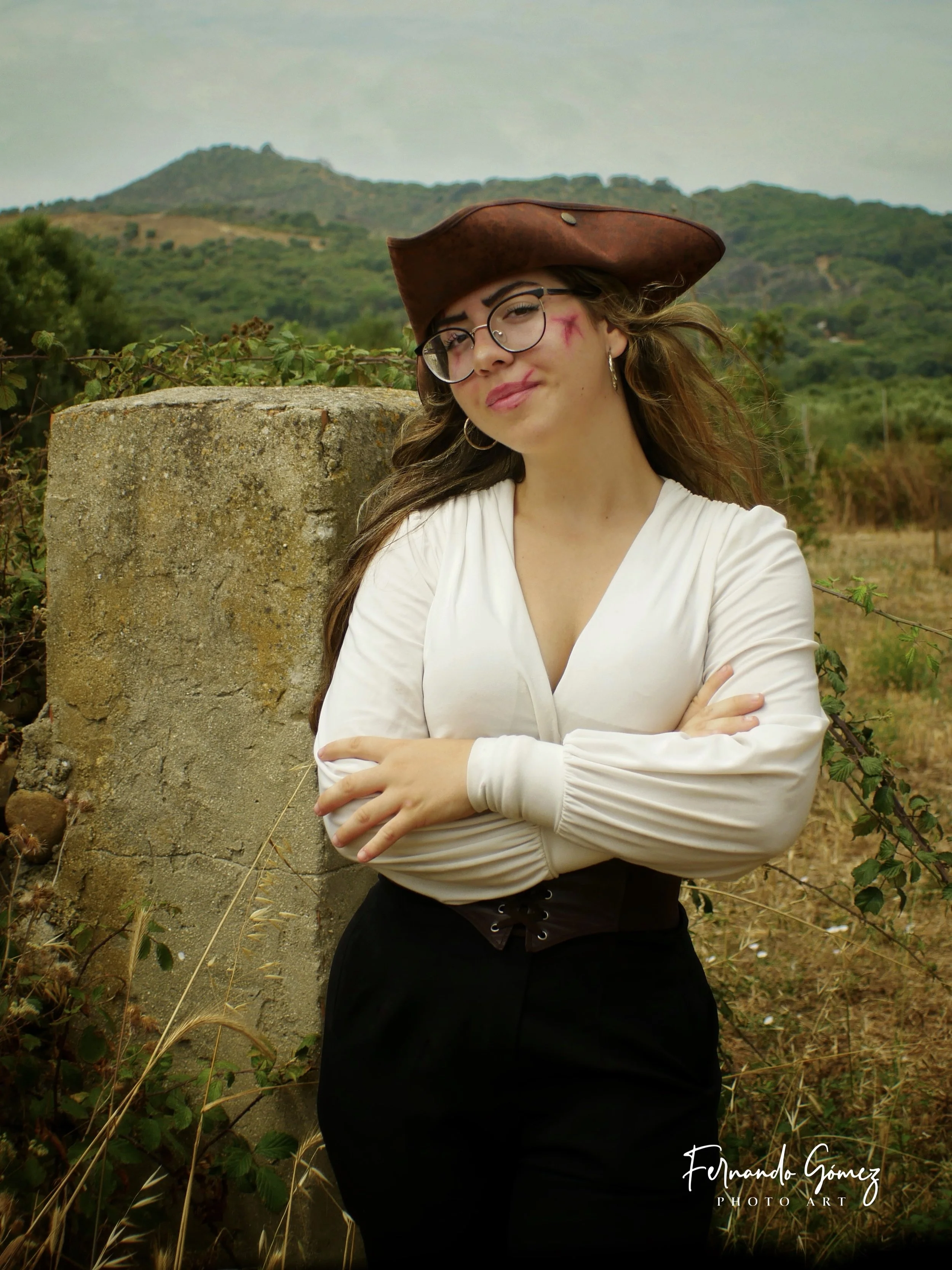 Young woman dressed in a white blouse and black high-waisted pants, wearing a brown tricorn hat and glasses, stands outdoors near a stone wall covered with greenery, with hills and trees in the background, and crosses her arms with a slight smile.