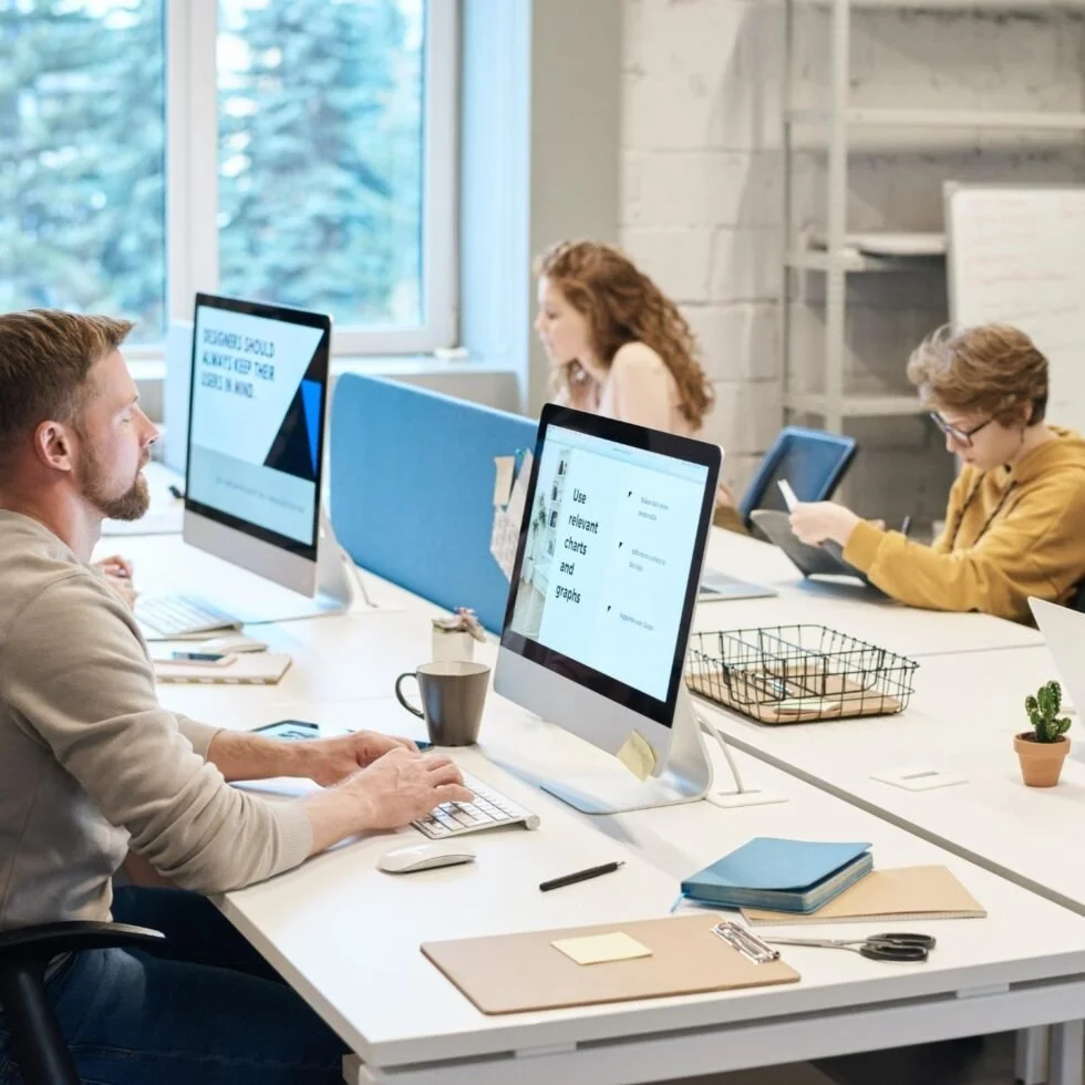 Tres personas trabajando en una oficina con computadoras, un hombre en primer plano, una mujer en el fondo y otra joven a la derecha, en un entorno moderno y luminoso.