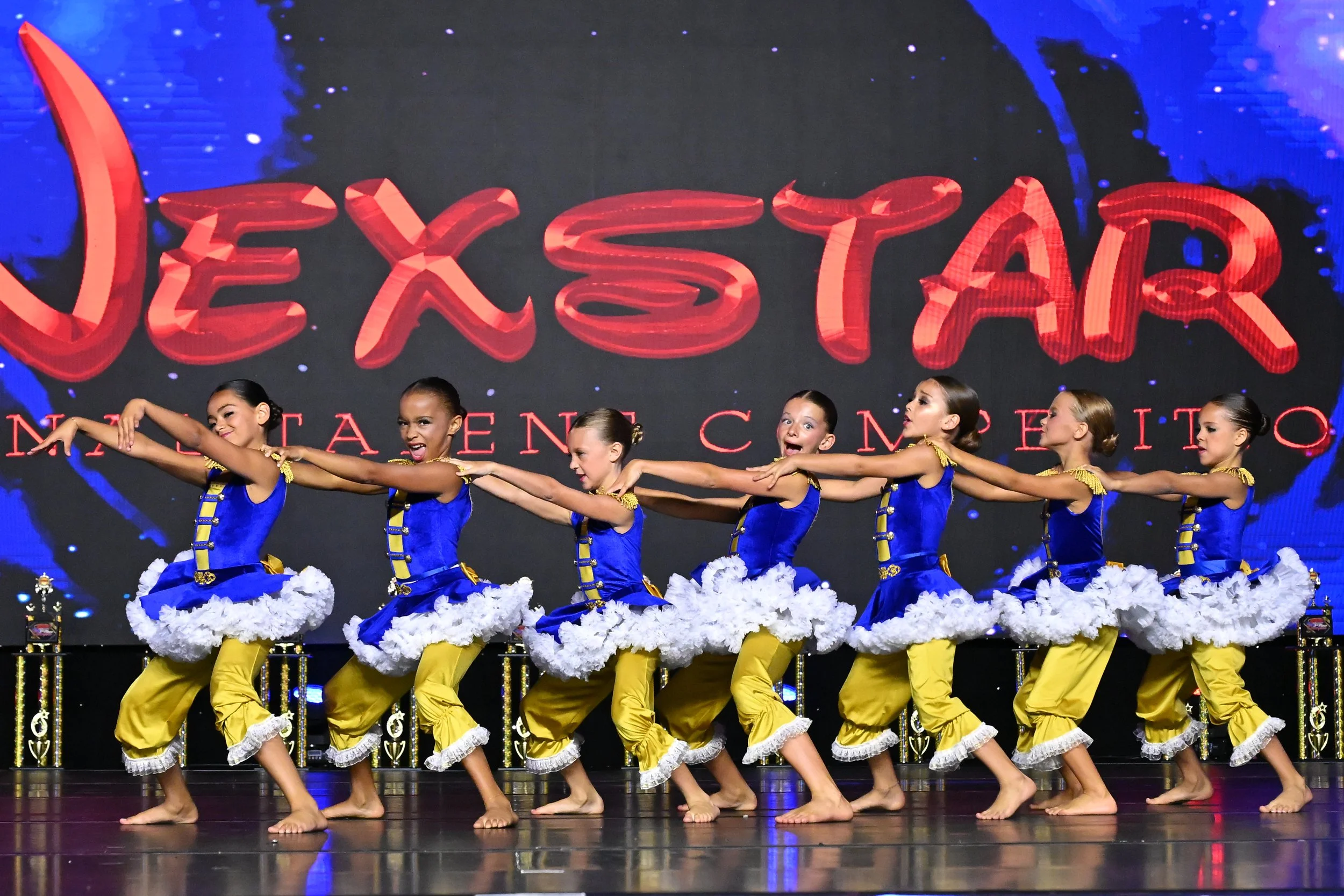Group of young girls in blue and yellow costumes performing a dance on stage with a large digital sign in the background reading 'EXSTAGE' and 'NATIONAL ENTERTAINMENT'.