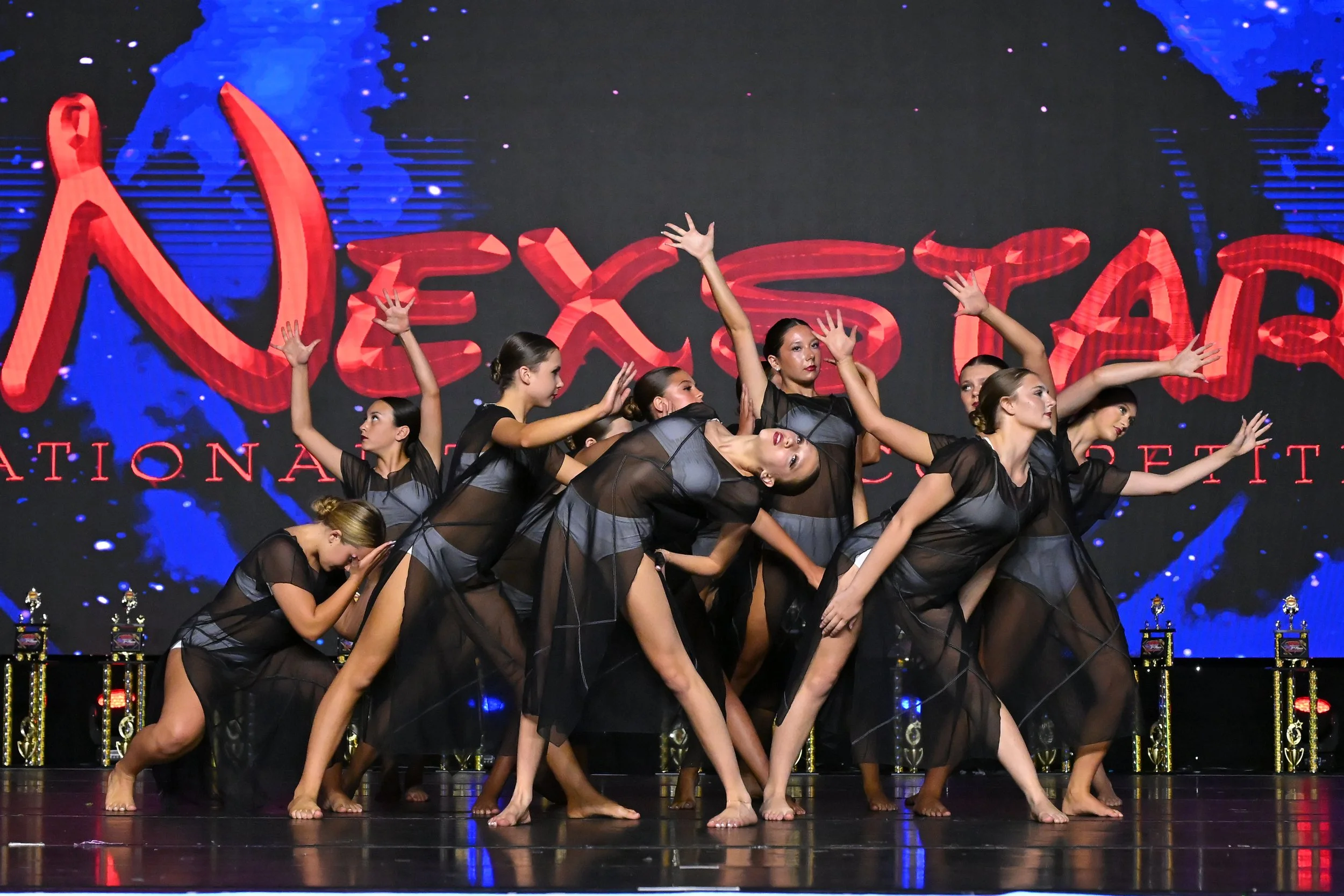 Young dancers performing on stage in black sheer costumes, backing a large screen displaying red text and blue background, with trophies lined up in front.