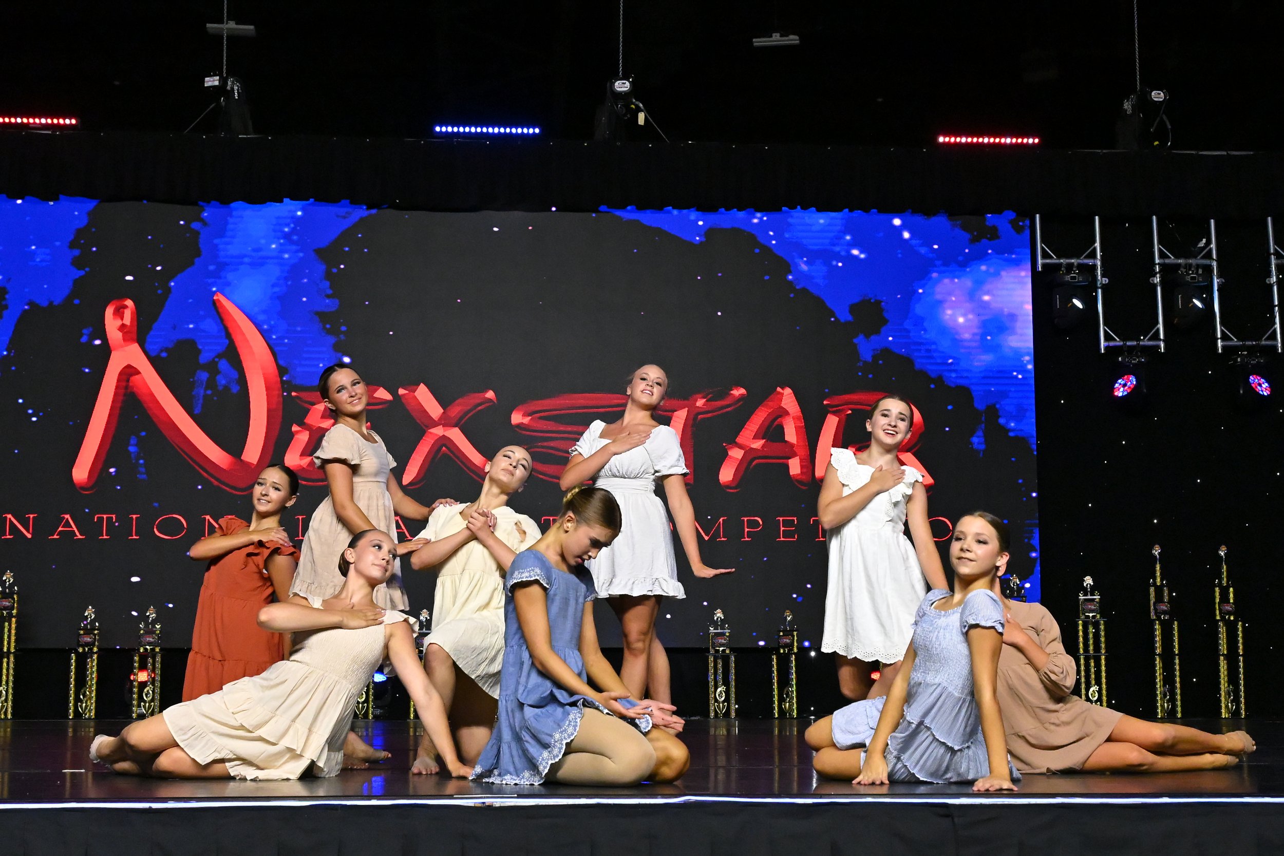 Young dancers in vintage-style dresses perform a choreographed dance on stage during a competition. The backdrop features a digital world map and the word 'Dance' in large red letters. Trophies are visible along the front of the stage.