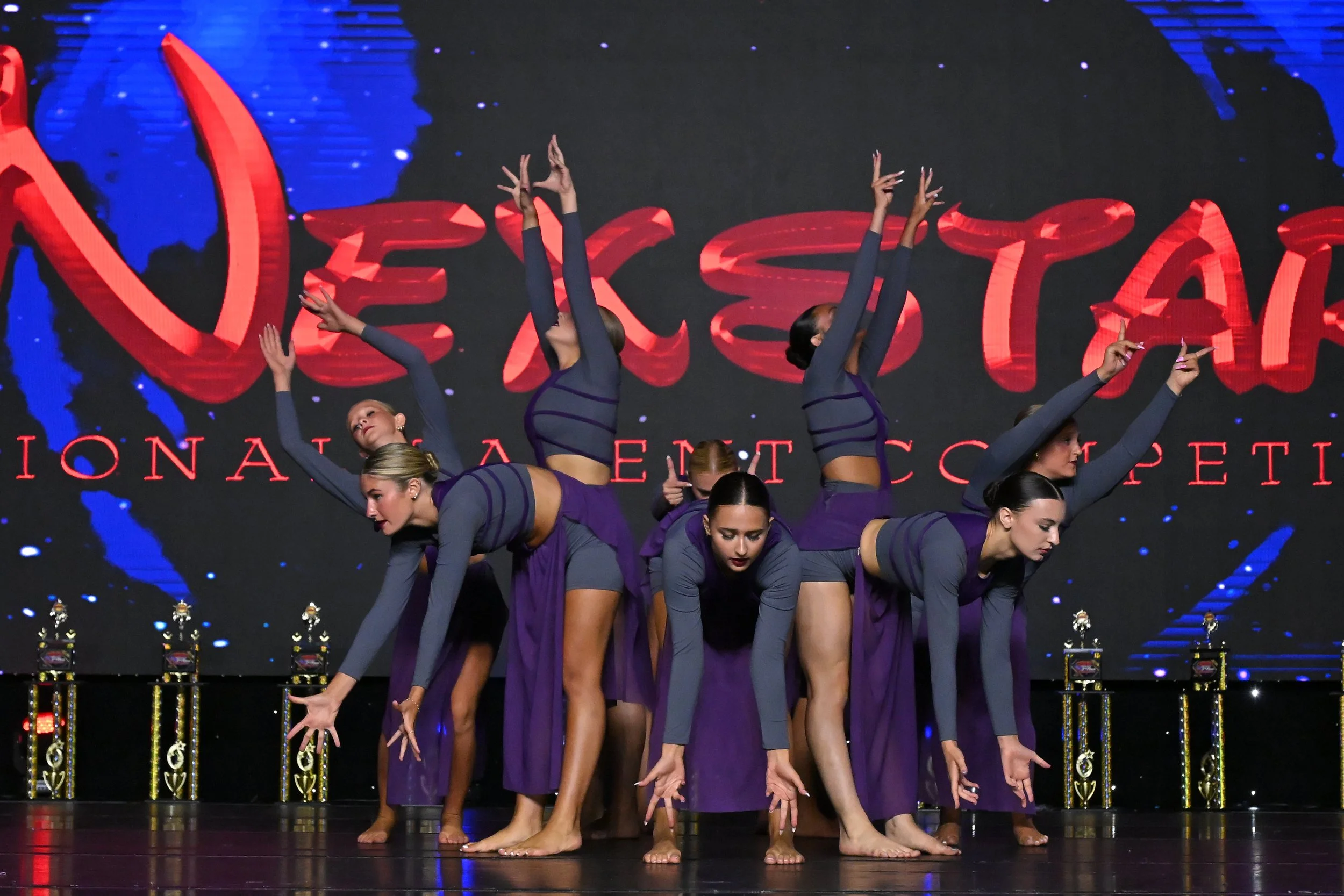 A group of dancers performing a choreographed pose on stage in matching purple and gray costumes with a large digital backdrop showing red and blue graphics behind them.