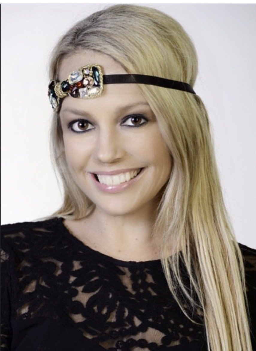 A woman with blonde hair wearing a black lace top and an ornate headband with colorful gemstones, smiling at the camera.