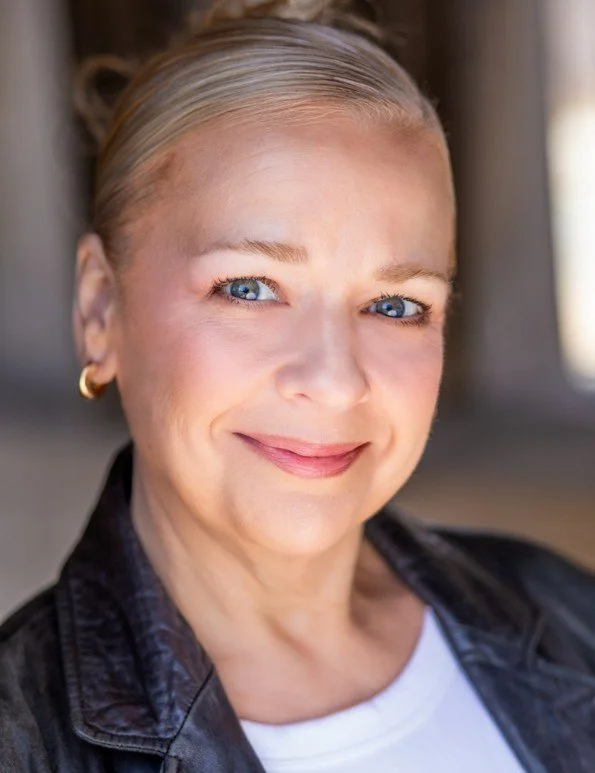 Close-up of a woman with blonde hair, blue eyes, wearing a black leather jacket, and gold hoop earrings, smiling outdoors.