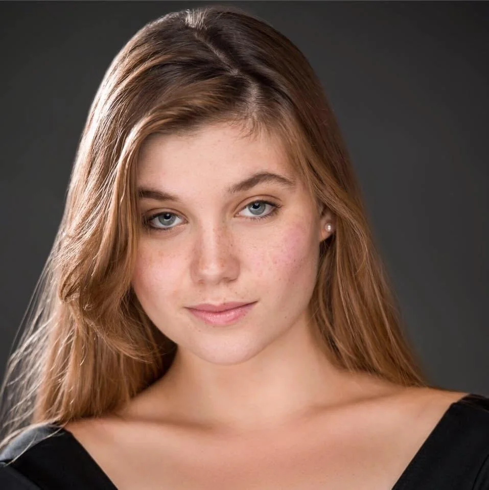 A young woman with fair skin, light blue eyes, and long, wavy, light brown hair looking directly at the camera on a dark background.