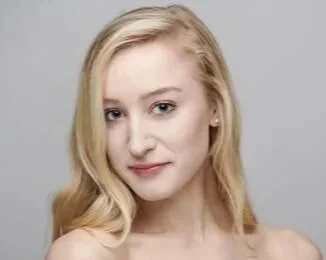 A young woman with long blonde hair looking at the camera with a slight smile, against a plain gray background.