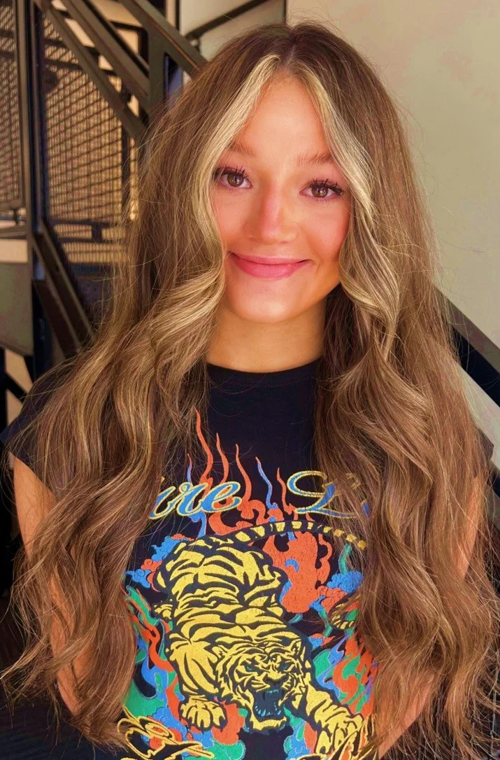 A young woman with long, wavy, light brown hair with blonde highlights, smiling, wearing a black T-shirt with a colorful graphic of a tiger and flames on the front, standing in front of a staircase with metal railings.