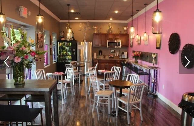 Interior of a brightly lit café or restaurant with pink walls, wooden floors, white chairs, and tables set for dining. There are hanging pendant lights, a small kitchenette in the background, and a flower arrangement on a table.