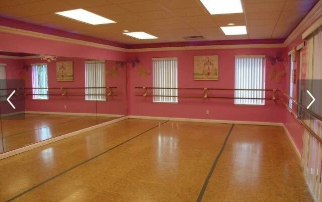 Empty dance studio with pink walls, three windows with vertical blinds, wooden floor, and wall-mounted ballet barres.