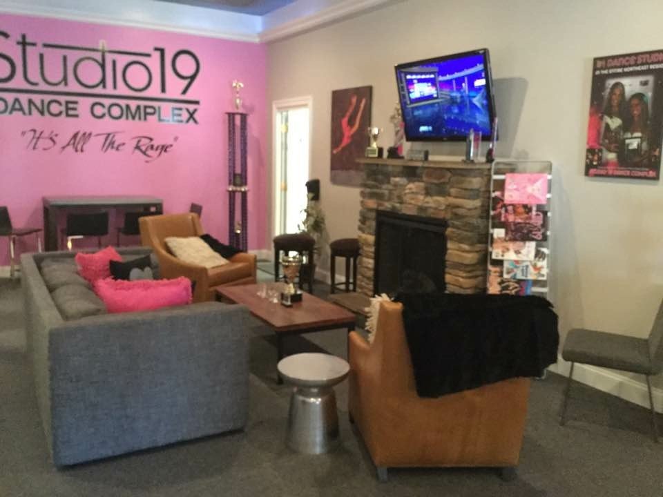 Indoor lounge area featuring a stone fireplace with a television mounted above, a pink wall with a sign reading 'studio19 DANCE COMPLEX' and 'It's All the Rage,' and various chairs and couches with pillows, along with a wall with framed posters and a