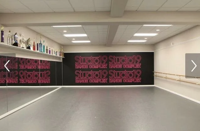 Empty dance studio with a black wall decorated with pink neon sign that reads 'Studio 19 Dance Complex', a mirror on the left wall, and a ballet barre on the right wall. Trophies are displayed on a shelf above the mirror.