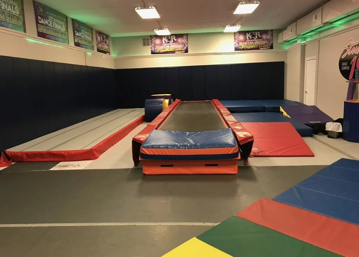 Empty indoor gymnastics training area with mats, trampolines, and foam blocks for tumbling and gymnastic practice, illuminated by ceiling lights with posters on the walls.