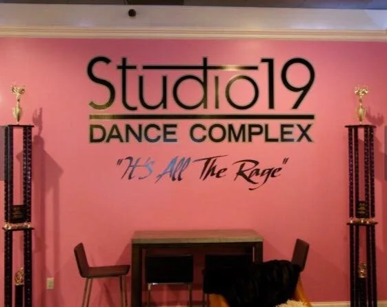 Pink wall with a Studio 19 Dance Complex sign and slogan 'It's All The Rage', surrounded by tall trophies on shelves.