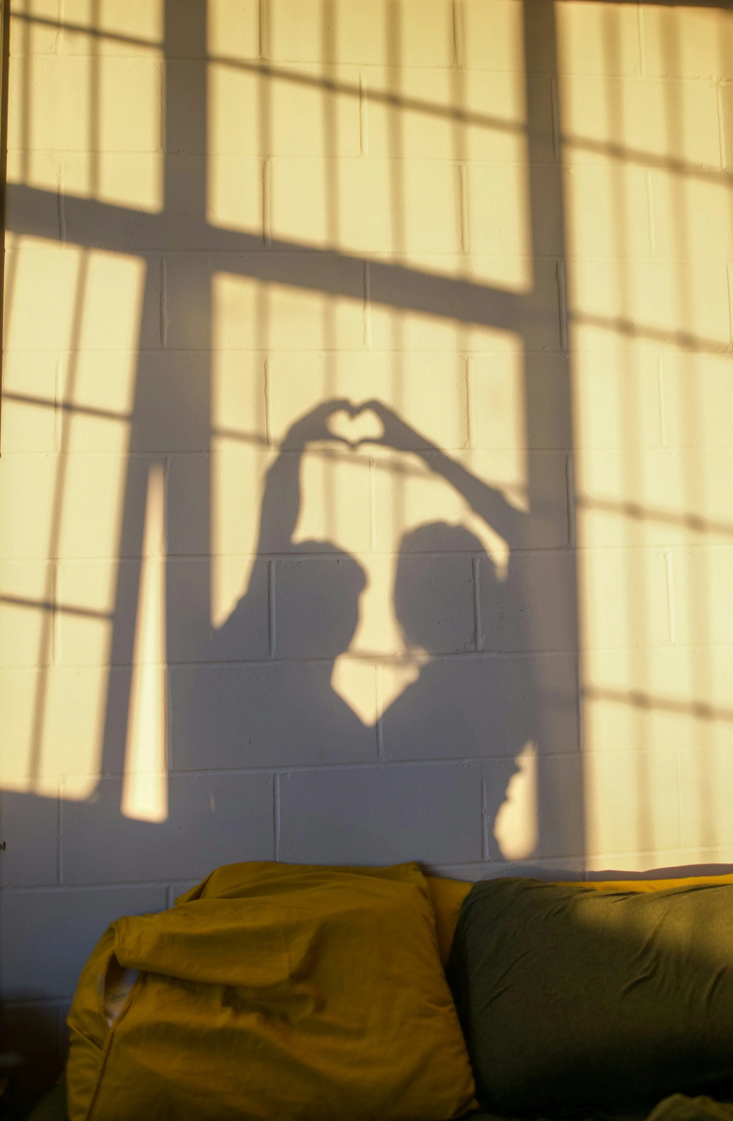 Sunlight creates the shadow of a person's hands forming a heart shape on a brick wall. There is a yellow pillow and a dark grey pillow on a bed or sofa in the foreground.