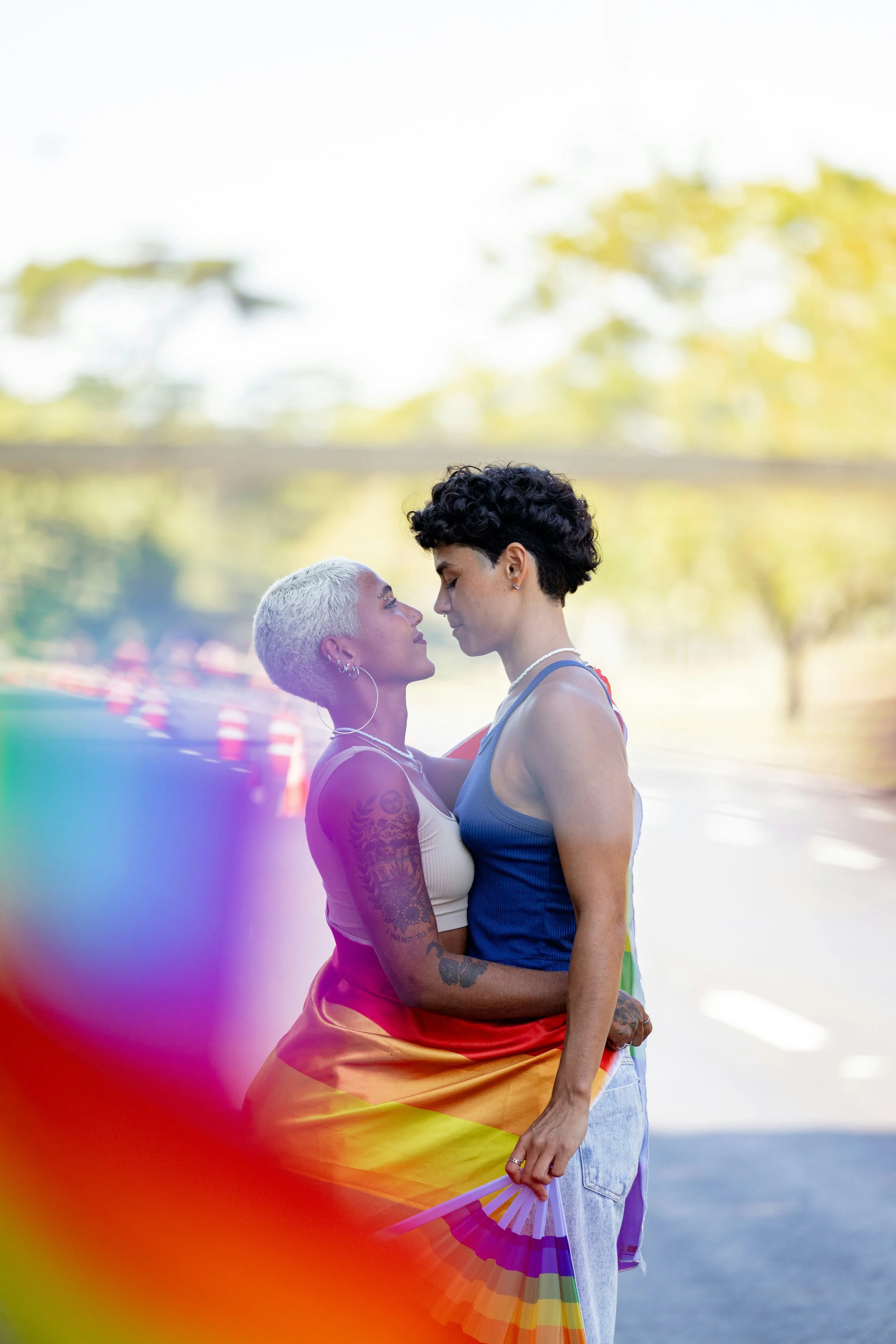 Two women standing close together outdoors, embracing and looking into each other's eyes, with a colorful rainbow flag wrapped around them.