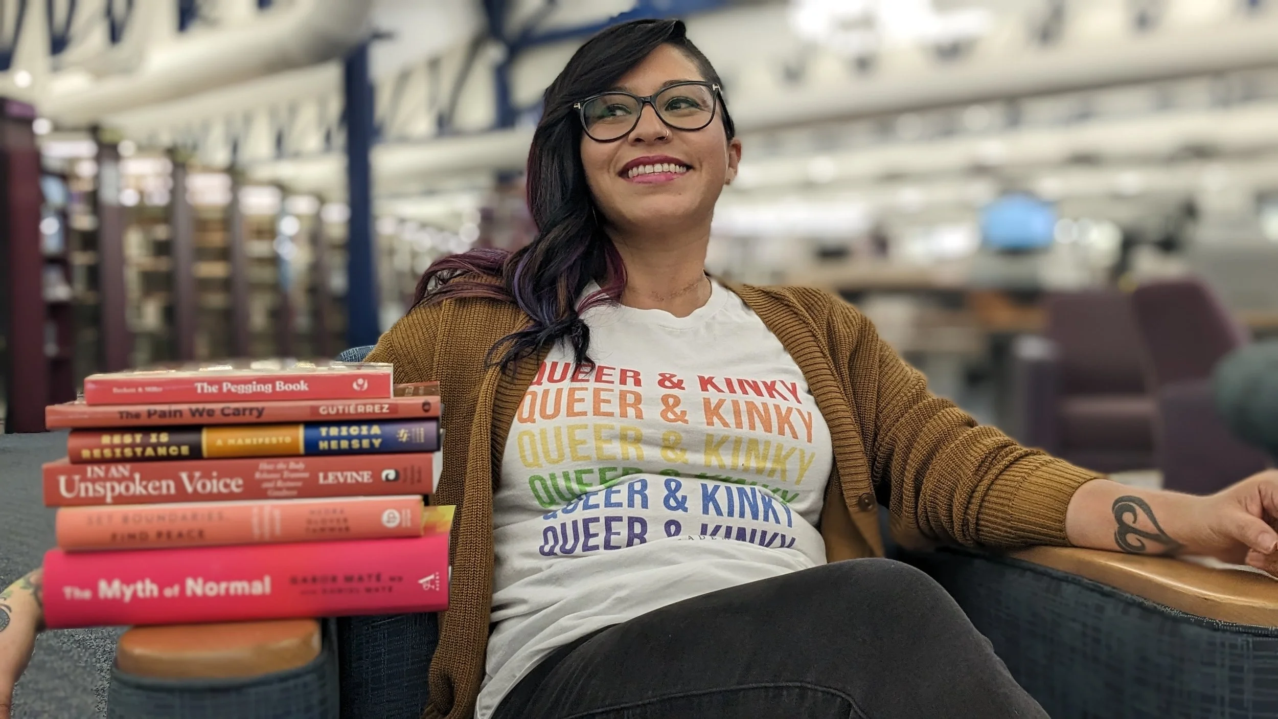 A woman with dark hair, glasses, and a rainbow-colored QUEER & KINKY t-shirt, smiling and sitting near a stack of books at an airport lounge or train station. The background features metal architecture and blurred seating.