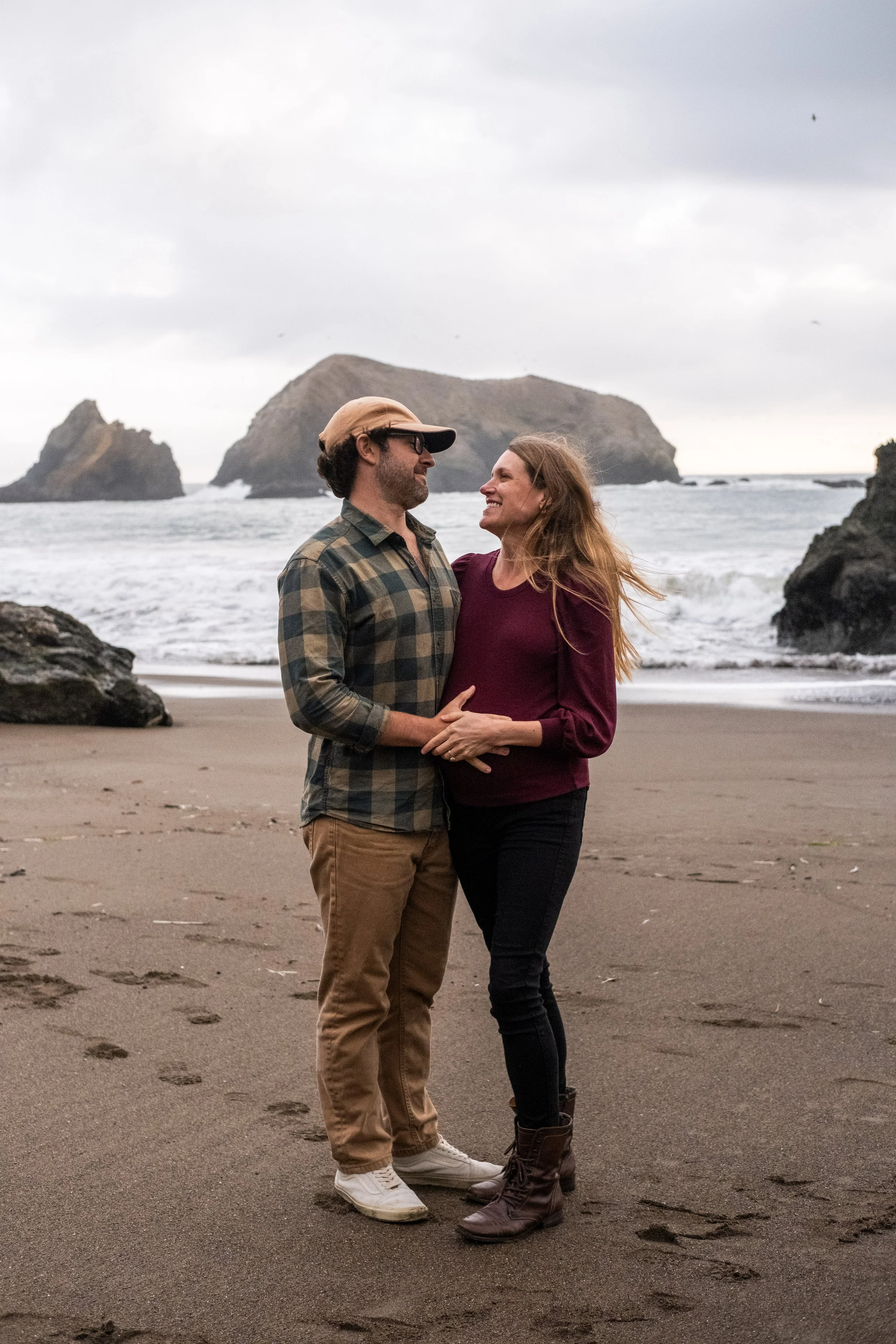 A man and pregnant woman standing on a beach, facing each other and smiling, with large rock formations and the ocean in the background, during a maternity photoshoot at Rodeo Beach.
