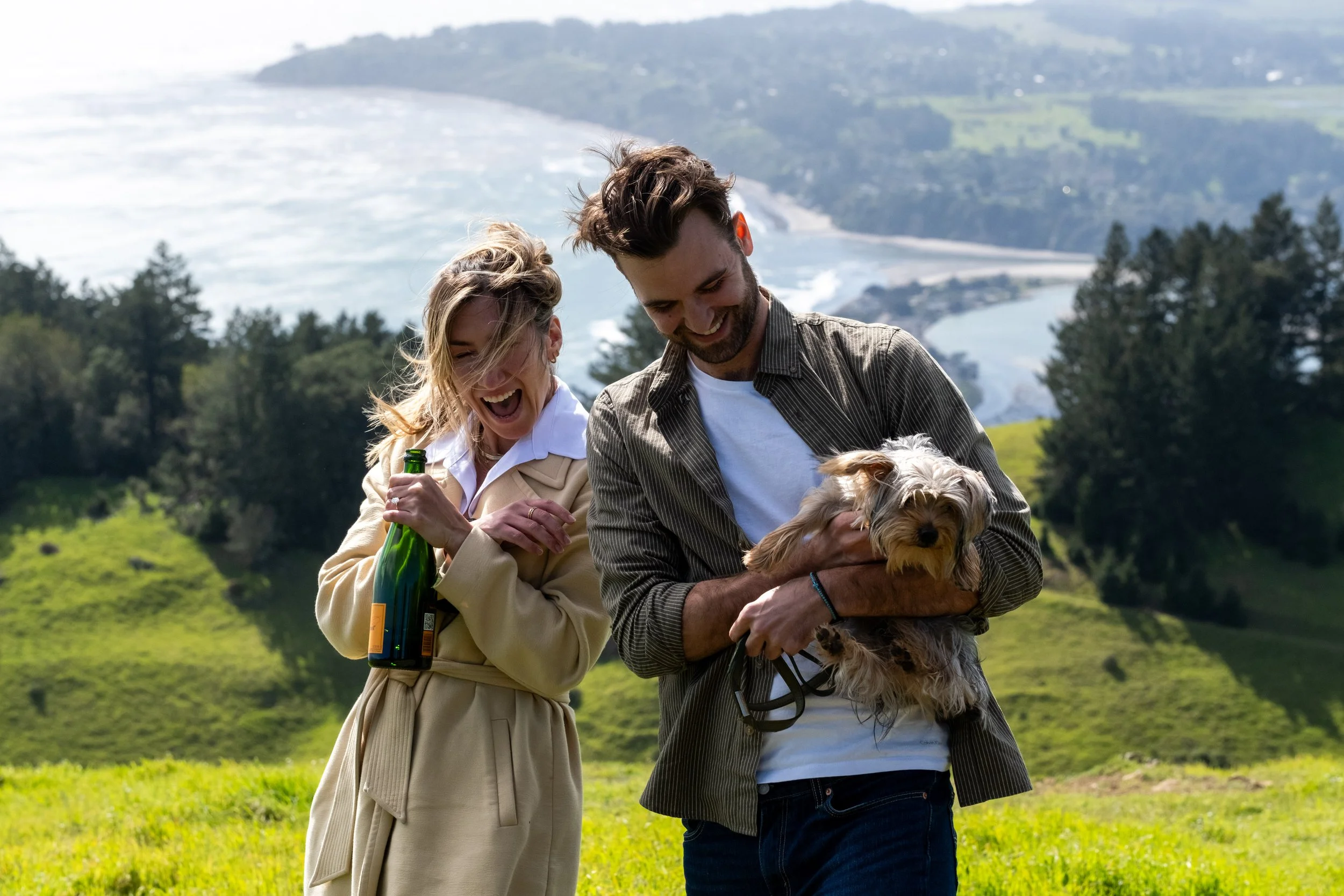 Two people, a woman and a man, smiling and looking at a small dog they are holding, outdoors on a grassy hillside with trees and a distant water body and hills in the background during an engagement photoshoot on Mt. Tamalpais.