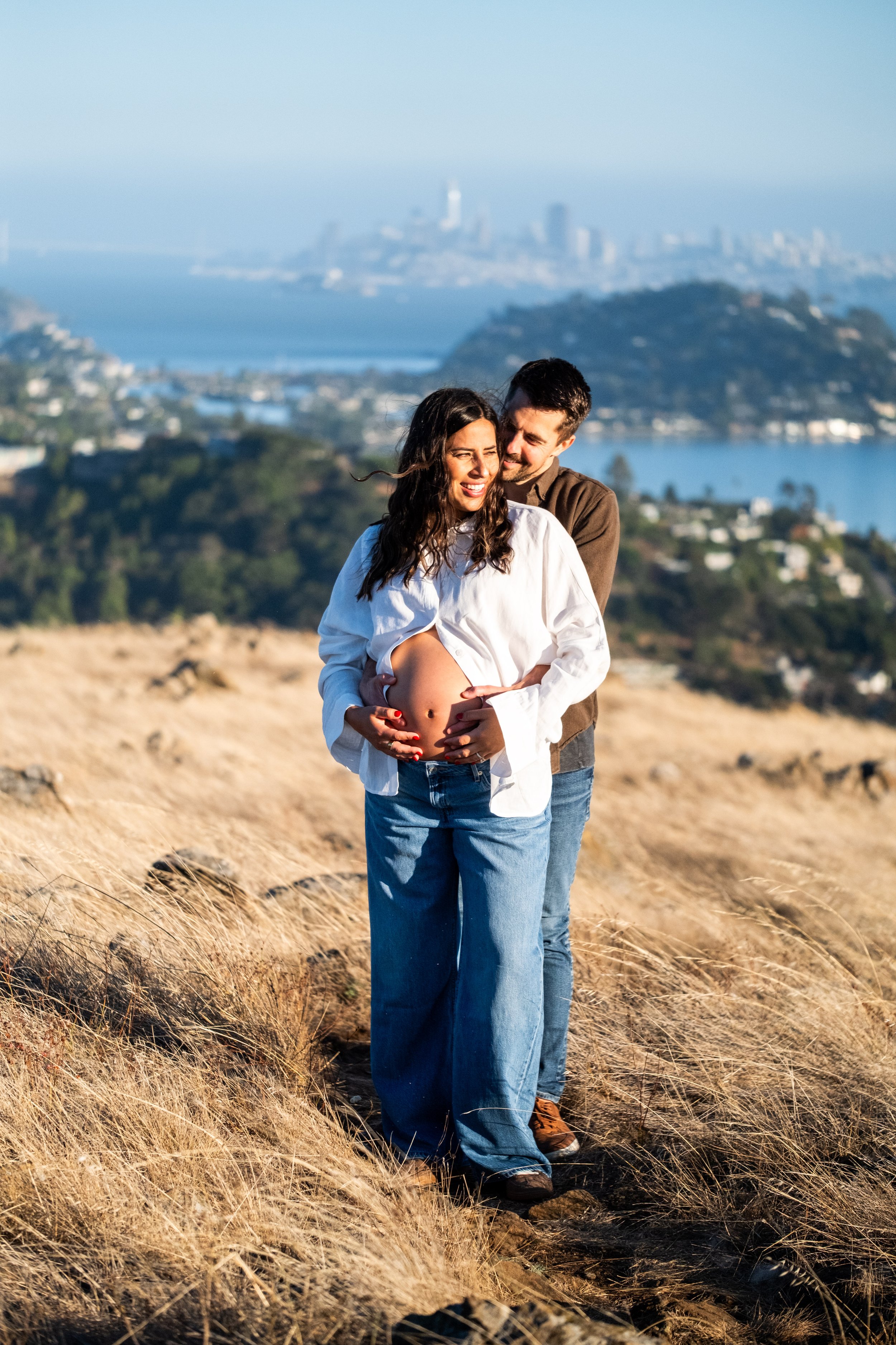 A pregnant woman and a man standing on a dirt path in a grassy field with a city skyline and bay in the background, during daytime, for a maternity photoshoot in nature.