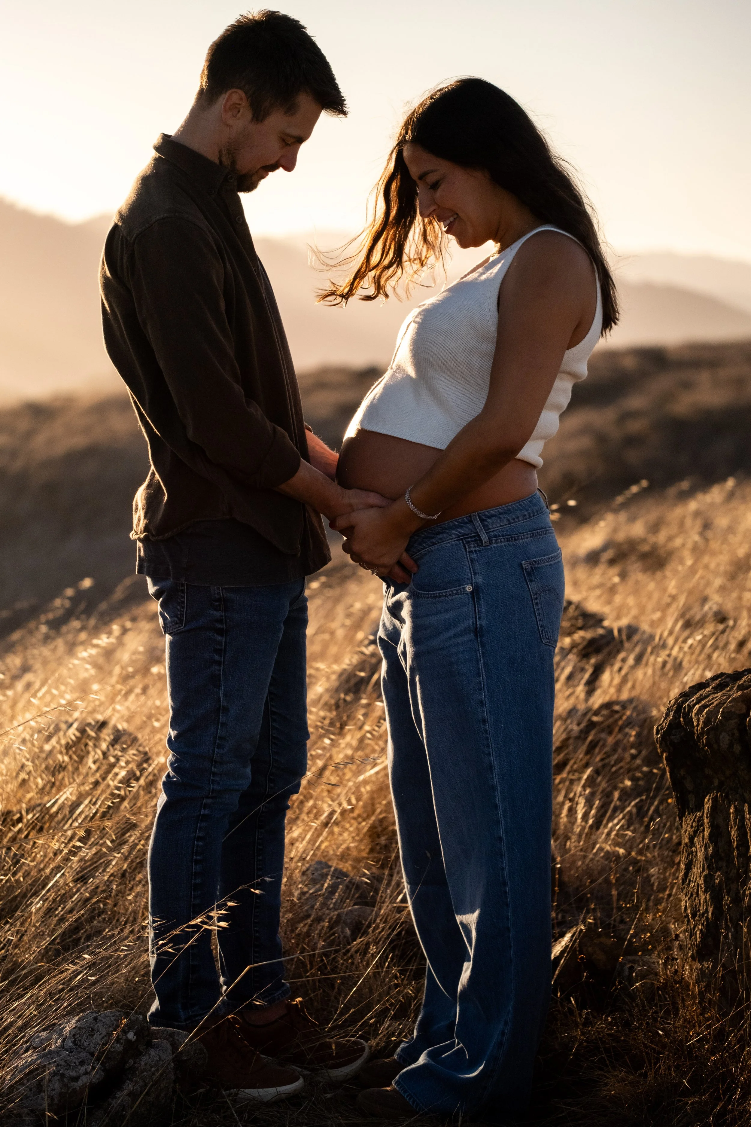 A couple stands close together outdoors during sunset, holding hands over a pregnant woman's belly, with rolling hills in the background, during a Marin County maternity photoshoot.