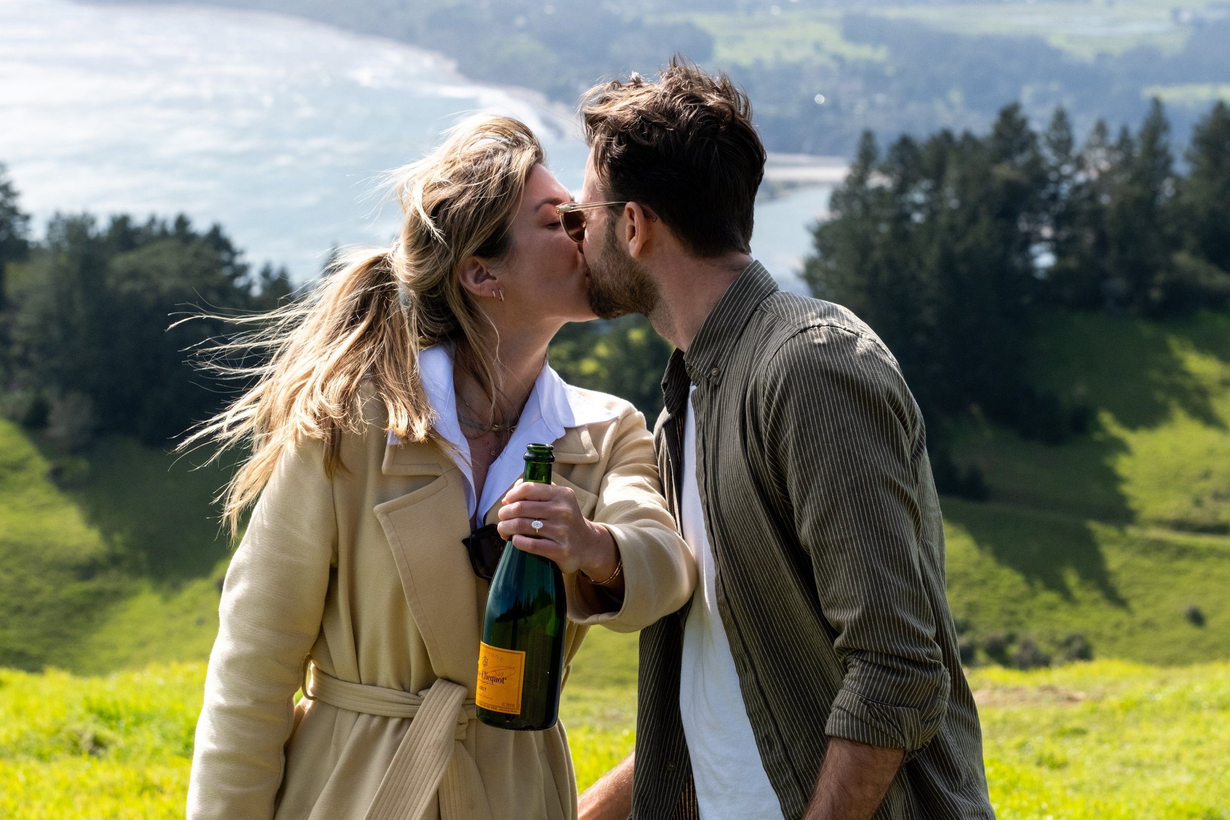 A couple kissing outdoors, with the woman holding a bottle of champagne, during a proposal photoshoot in Mill Valley.