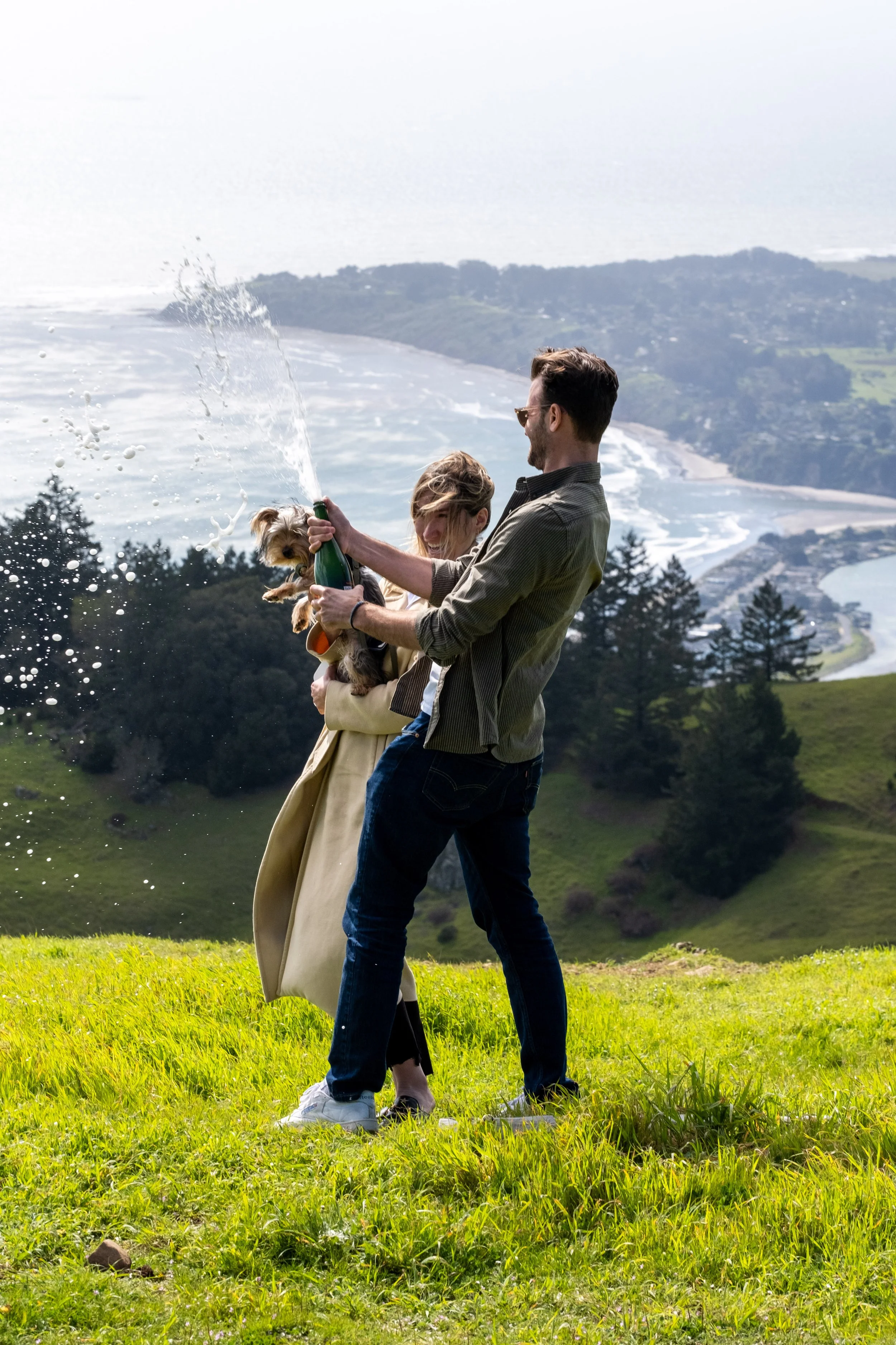 A man and woman are celebrating outdoors on a grassy hill, with the woman holding a small dog and the man opening a bottle, possibly champagne, in a scenic coastal area with mountains and the ocean in the background.