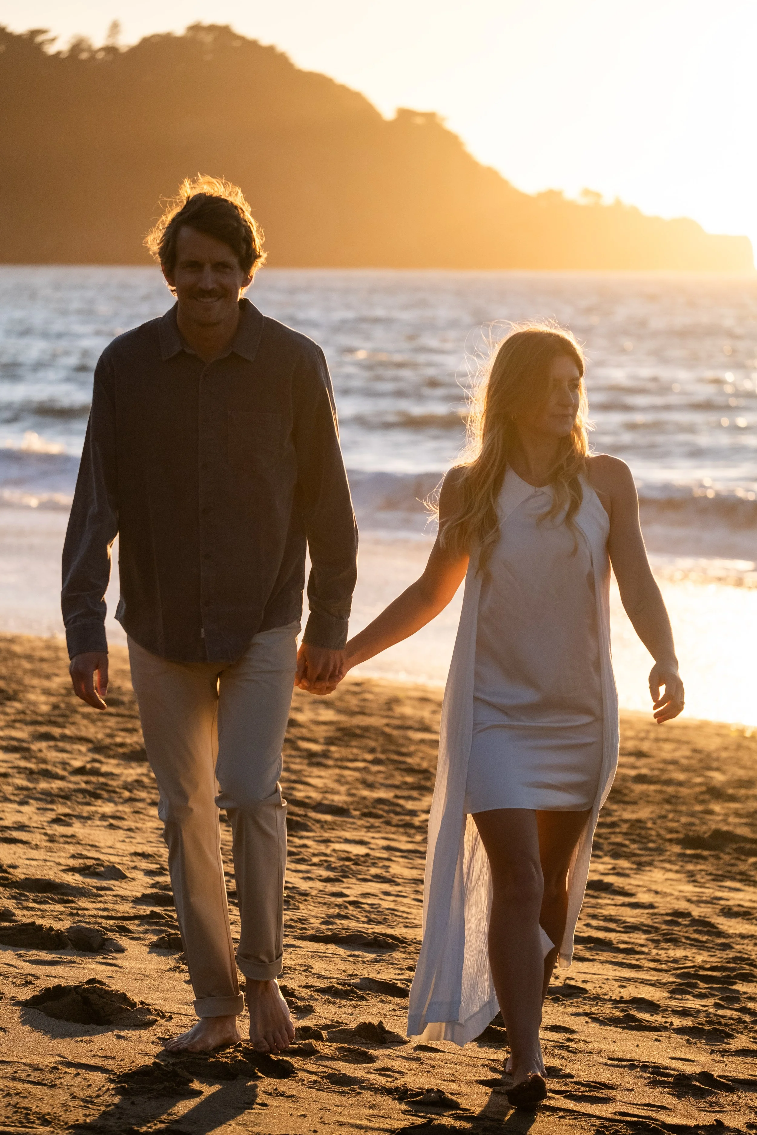 A man and a woman walking hand in hand on the beach during sunset, with the ocean and cliffs in the background during an engagement photoshoot in San Francisco.